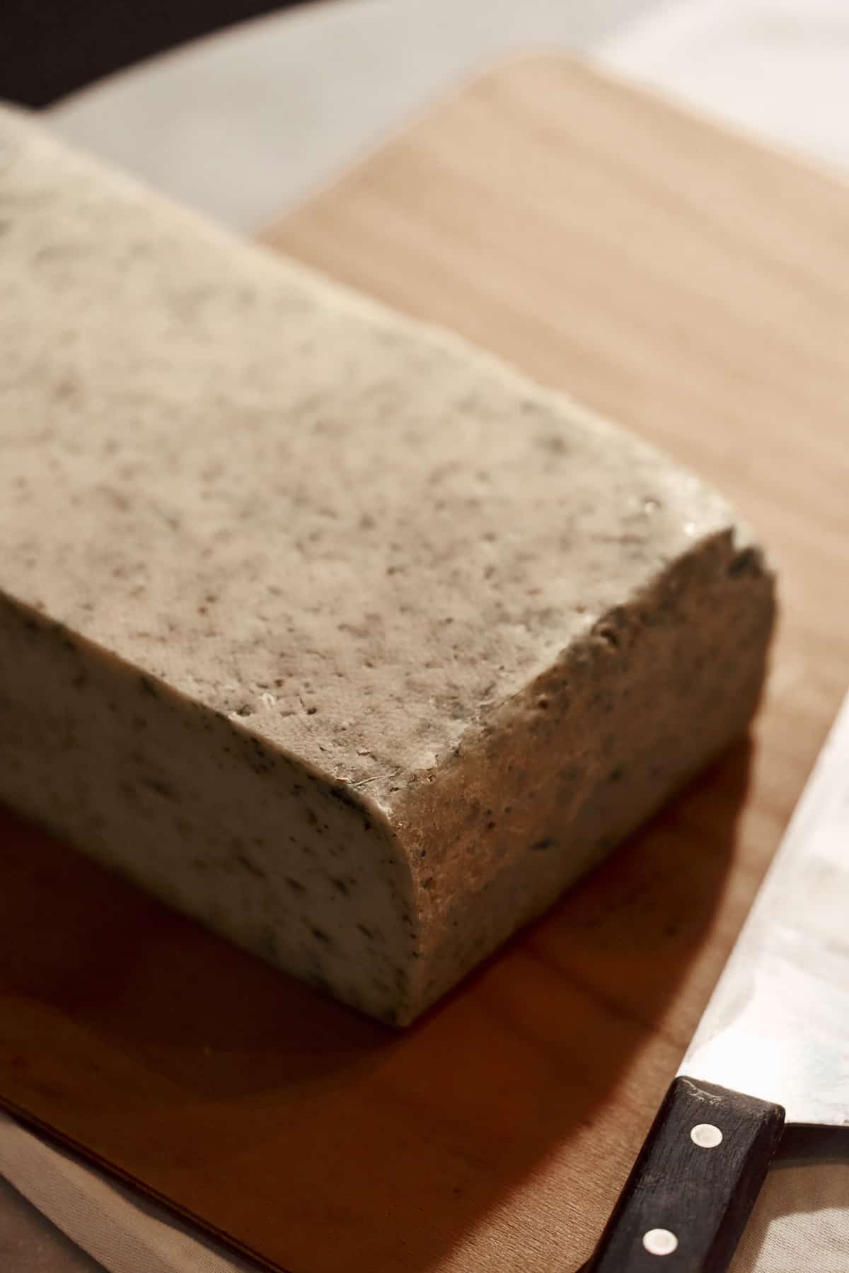 Ein rechteckiger Käselaib mit kräuterdurchsetzter Rinde steht auf einem Holzbrett. Im unscharfen Hintergrund ein Weinglas mit gefalteter weißer Stoffserviette und eine Stuhllehne.