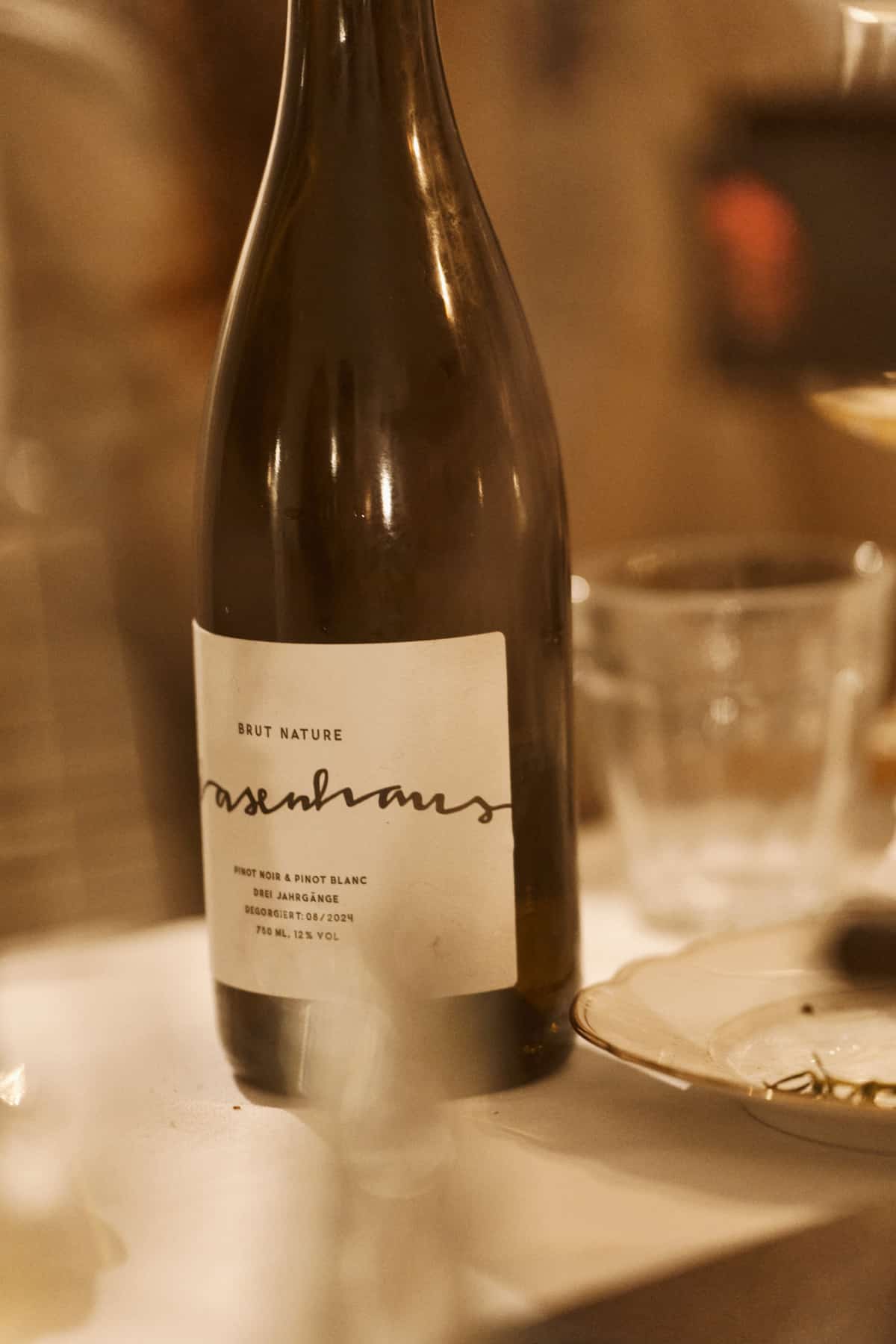 Nahaufnahme einer Flasche Wasenhaus Brut Nature (Pinot Noir und Pinot Blanc) mit hellem Etikett, im unscharfen Hintergrund ein Trinkglas und ein Teller.
