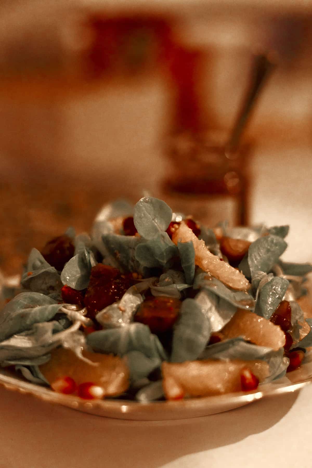 Teller mit einem Blattsalat aus grünen Blättern, dunklen Fruchtstücken und vereinzelten roten Kernen, im unscharfen Hintergrund ein Gefäß mit Besteck.