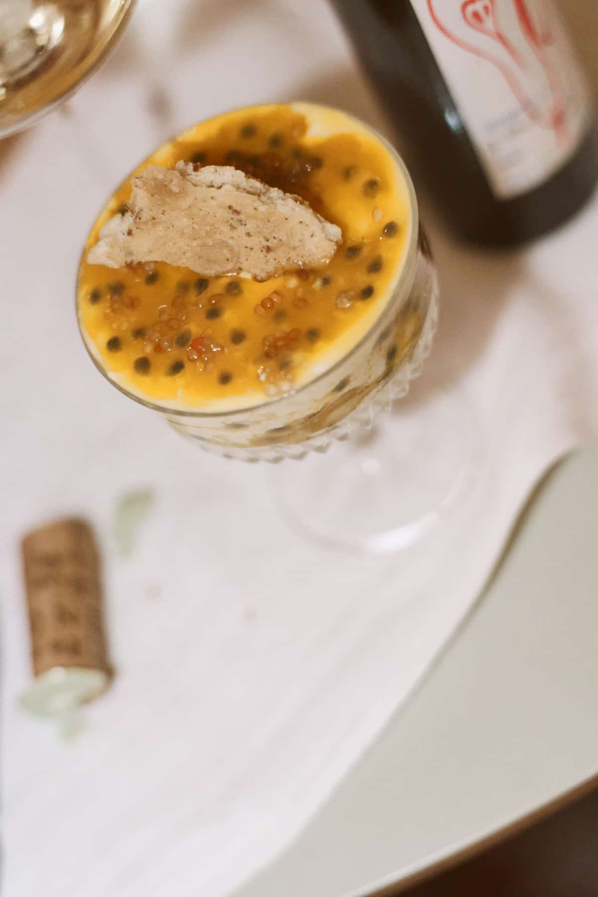 Dessert in einem facettierten Glas mit Maracuja-Sauce, einer Karamell-Tuile und Fruchtkernen, im unscharfen Hintergrund eine dunkle Flasche mit dem Etikett Obstler.
