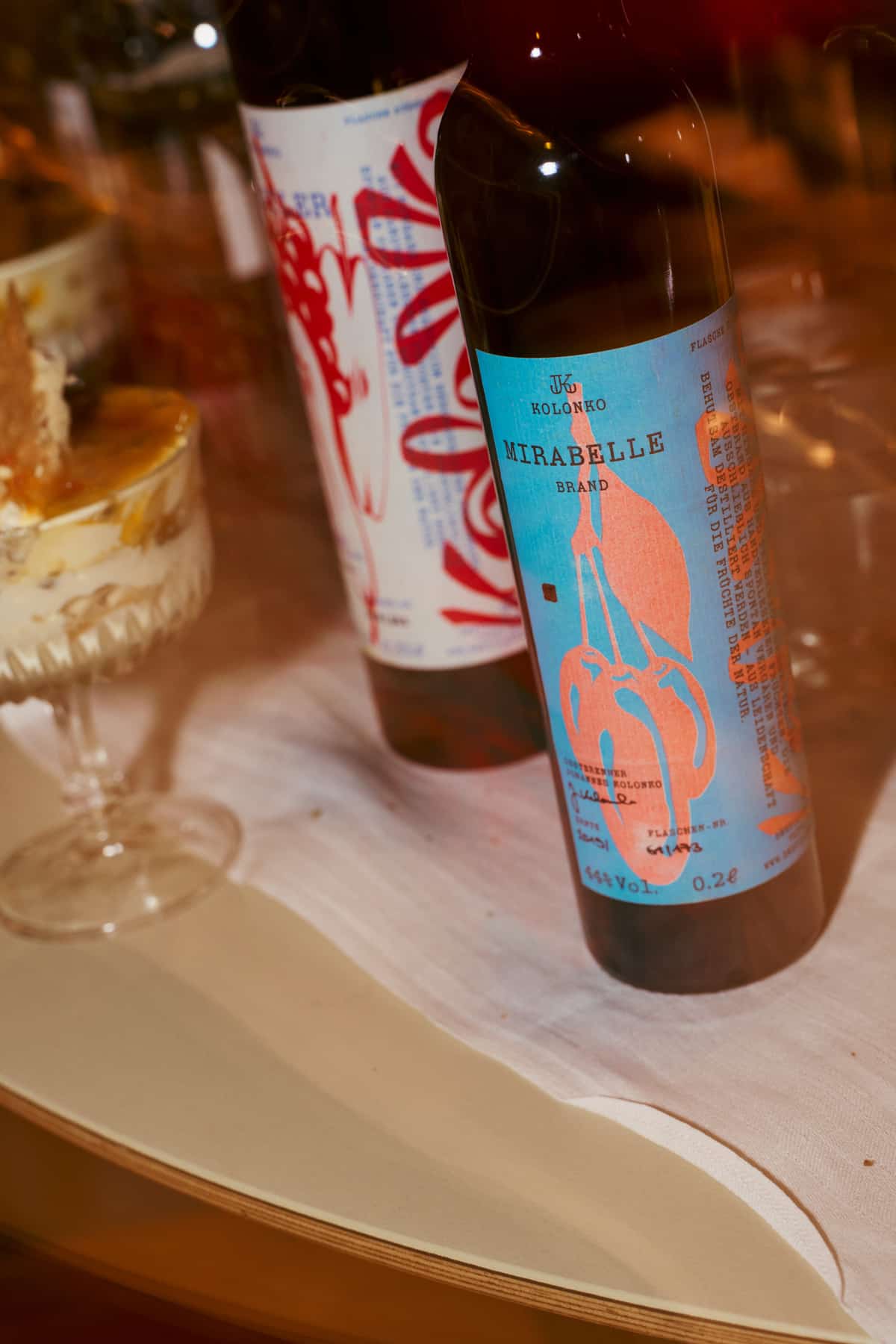 Nahaufnahme einer Spirituosenflasche mit blau-orangem Etikett Kolonko Mirabelle Brand, im unscharfen Hintergrund ein weiteres Flaschenetikett und ein Dessertglas.