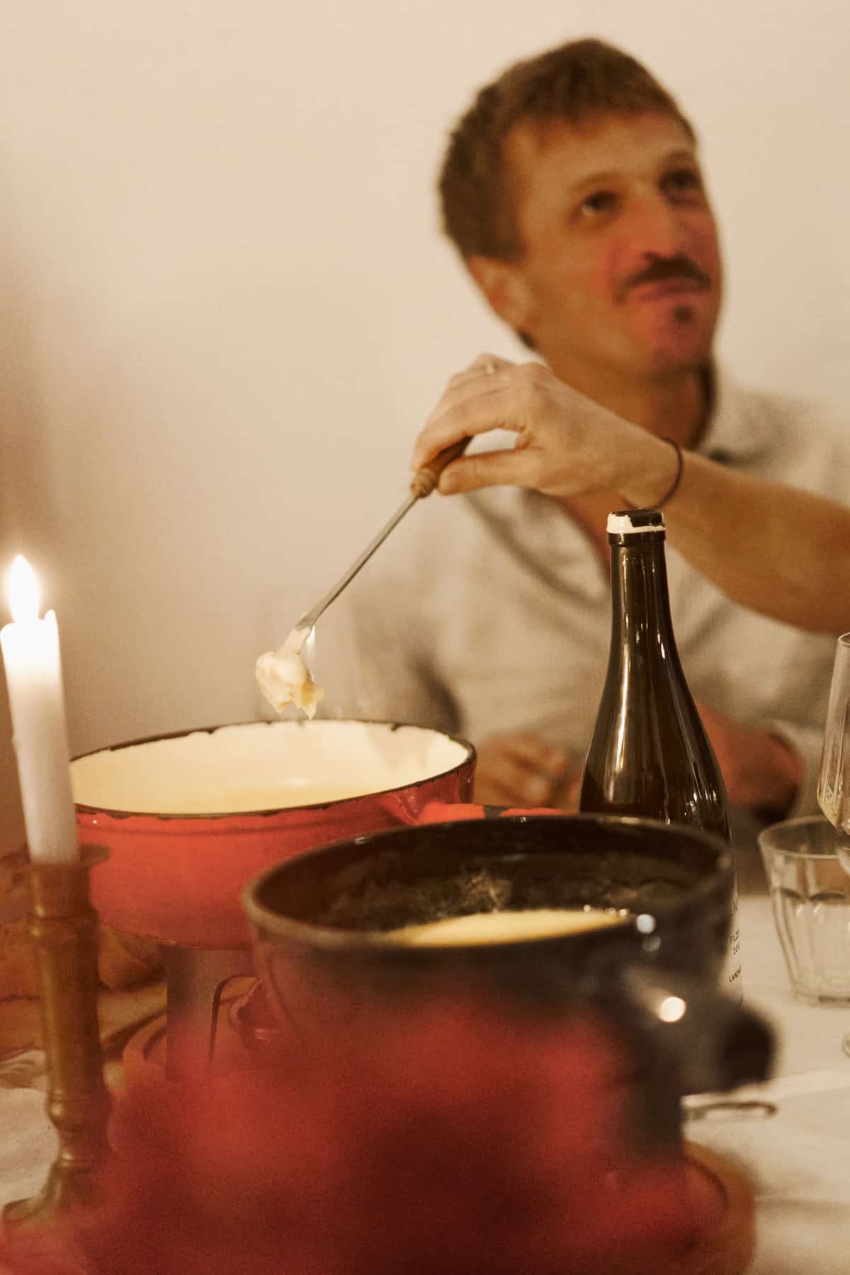 Ein Mann mit Oberlippenbart in gestreiftem Hemd taucht am Kerzenlicht eine Fonduegabel mit geschmolzenem Käse in einen roten und einen dunklen Fonduetopf, im Hintergrund eine Weinflasche und Trinkgläser.
