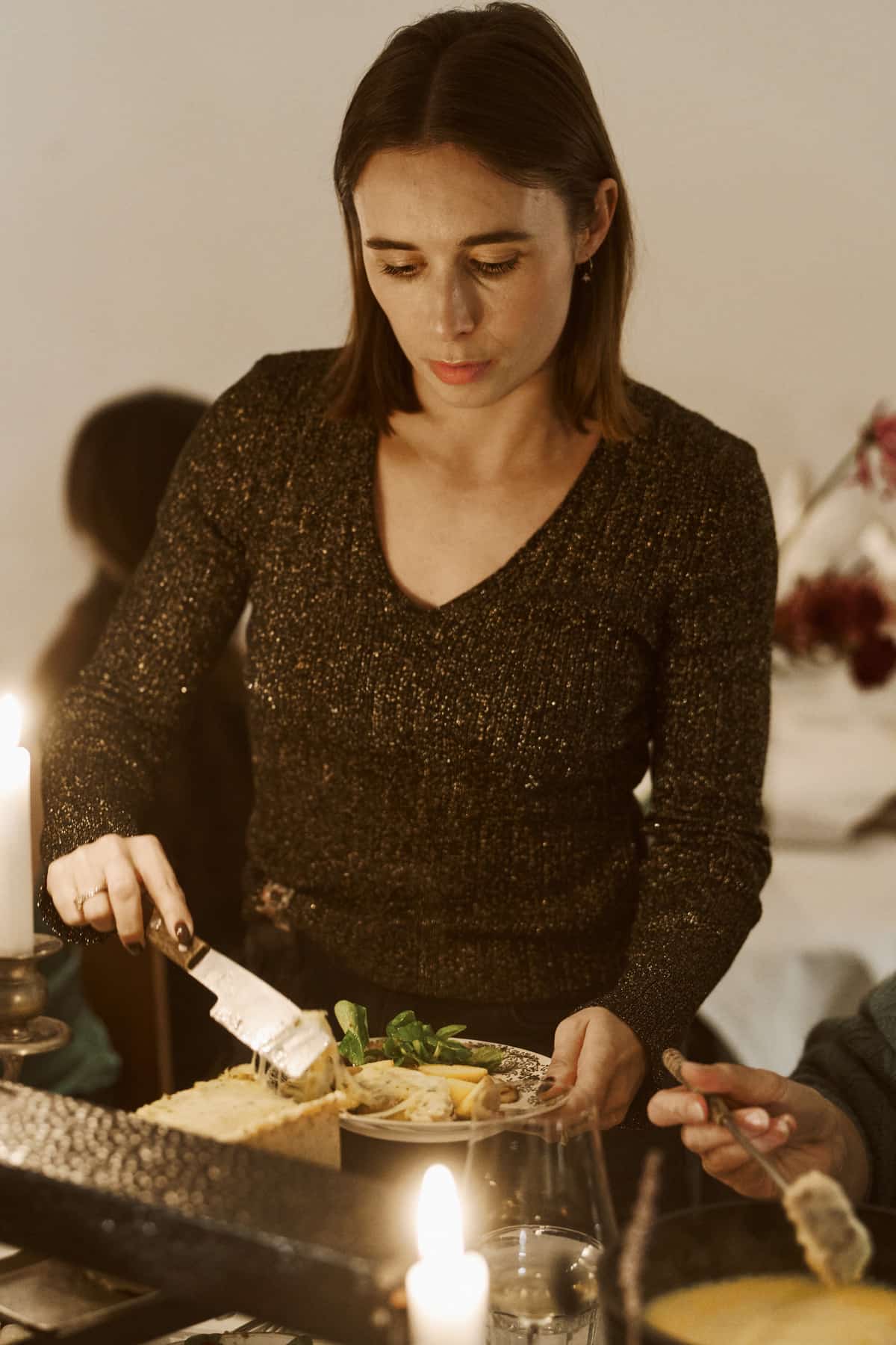 Eine Frau in glitzerndem dunklem Pullover schneidet konzentriert mit einem Käsemesser geschmolzenen Raclette-Käse auf einen Teller mit Blattsalat, an ihrem Handgelenk trägt sie einen Ring, im Vordergrund reicht eine zweite Person von rechts eine Käsestange auf einem Spieß heran.