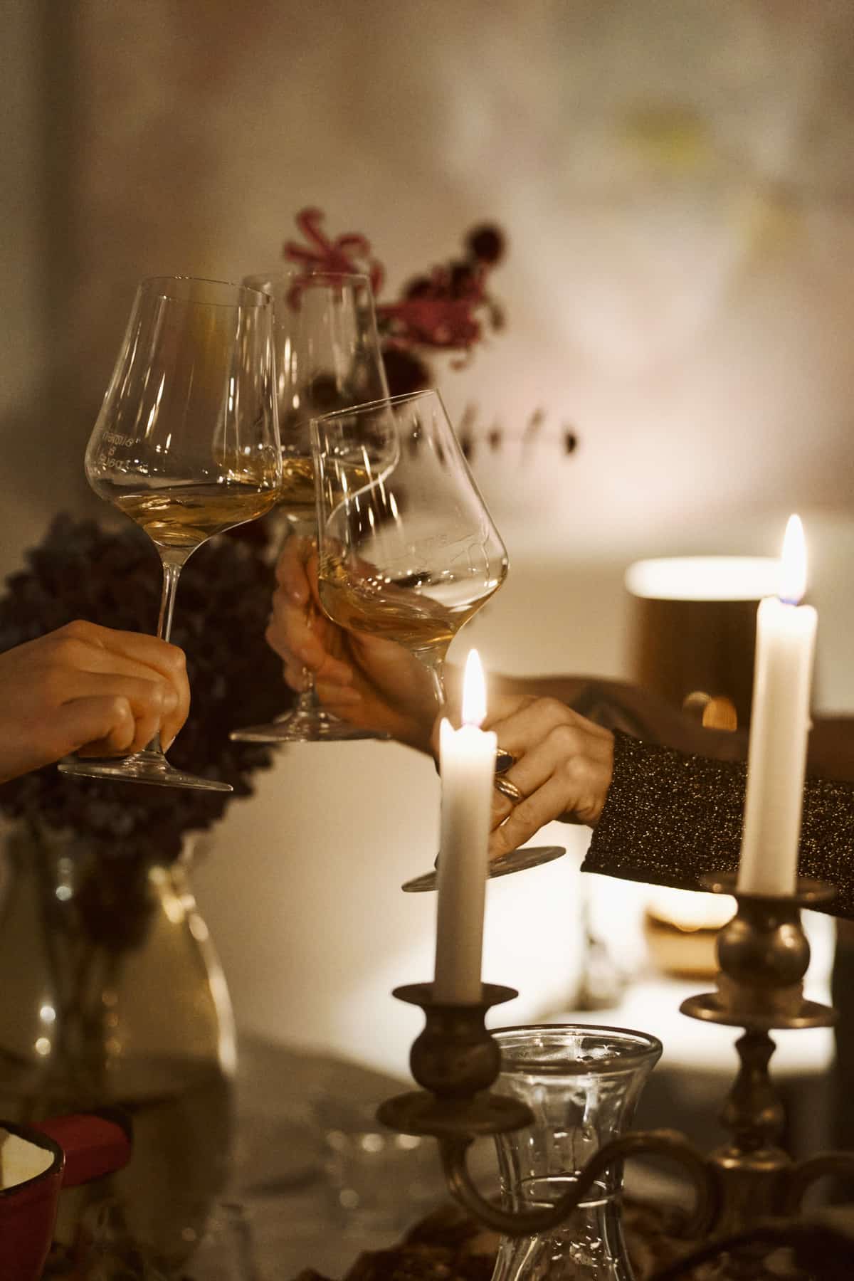 Drei Hände stoßen mit gefüllten Weißweingläsern über zwei brennenden Kerzen auf einem gedeckten Tisch an, im Hintergrund unscharf ein Blumenstrauß, keine Gesichter im Bild sichtbar.