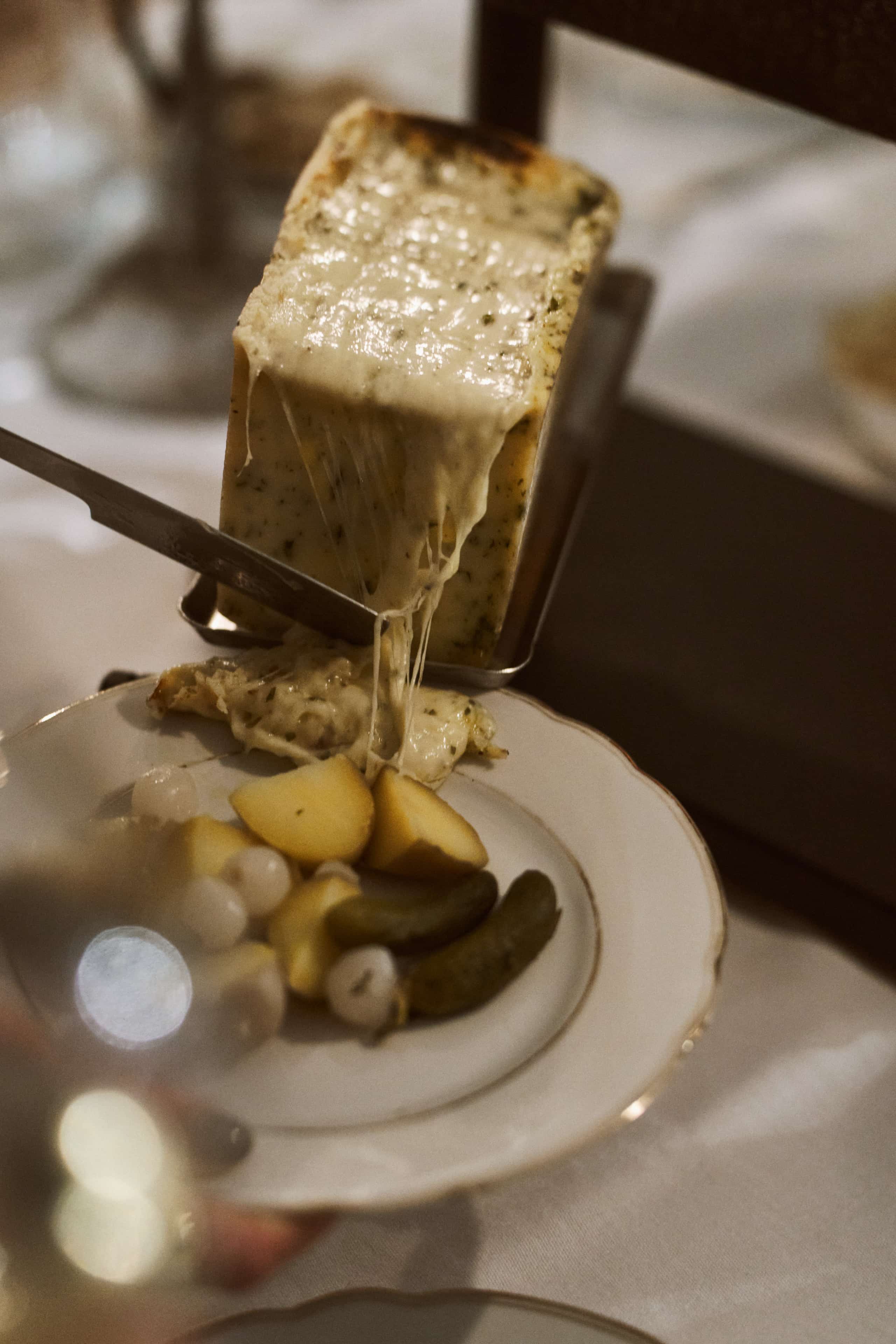 Nahaufnahme eines rechteckigen Raclette-Käselaibs unter einem Raclette-Grillelement. Die obere Schicht des Käses ist geschmolzen und läuft in Fäden auf einen darunter stehenden Teller mit Kartoffeln, sauren Gurken und Zwiebeln.