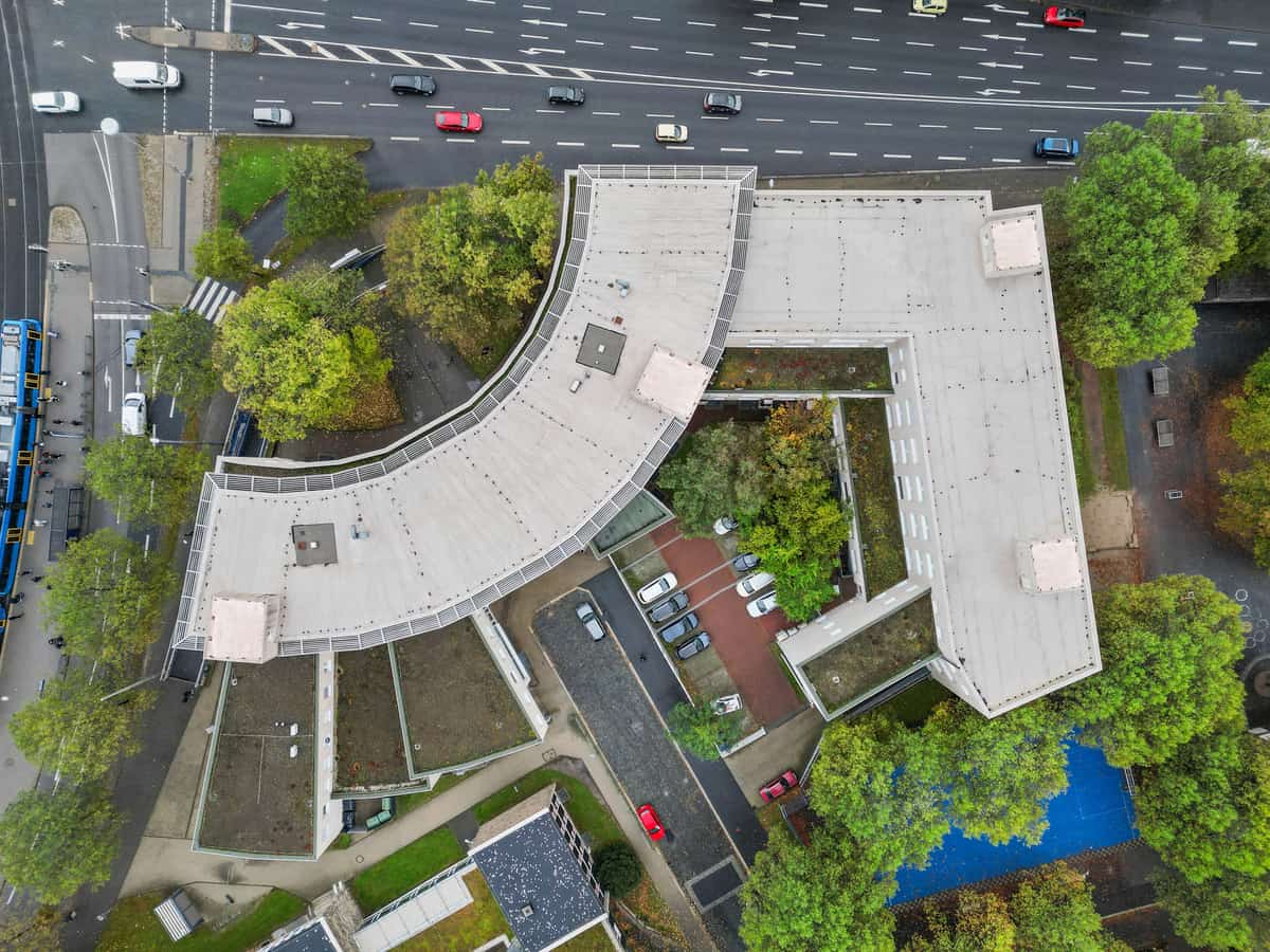 Luftaufnahme aus fast senkrechter Perspektive, die die geschwungene Grundrissform des Gebäudekomplexes mit Flachdach zeigt. Angrenzend eine Straße mit Gleisen und Baumreihen.
