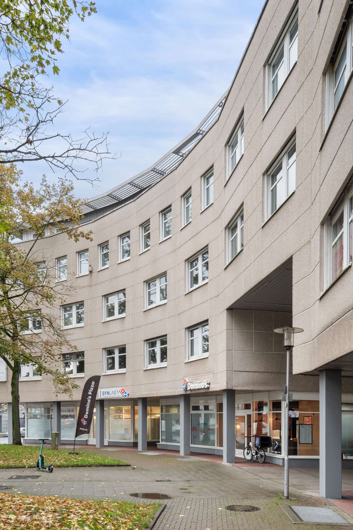 Straßenansicht der Gebäudefassade im Hochformat mit Gewerbeeinheiten im Erdgeschoss, darunter ein Domino’s-Pizza-Schriftzug und ein Synlab-Schriftzug. Im Vordergrund ein herbstlich gefärbter Baum, am Straßenrand geparkte Autos.