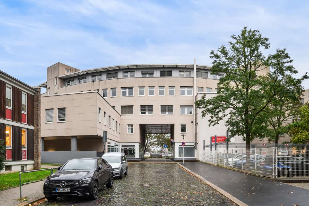Straßenansicht der Fassade mit einem verglasten Verbindungssteg zwischen zwei Gebäudeteilen. Ein Schild mit der Aufschrift Privat Parkplatz ist zu sehen, davor parkende Fahrzeuge und ein Baum.