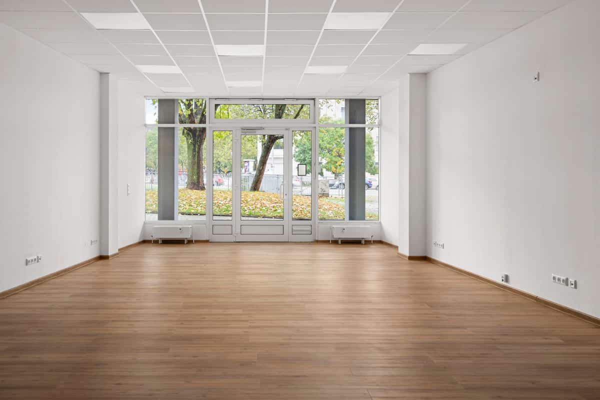 Innenansicht einer leeren Gewerbefläche im Erdgeschoss mit hellem Holzboden und einer großen Schaufensterfront zur Straße, durch die Bäume und parkende Autos draußen sichtbar sind.