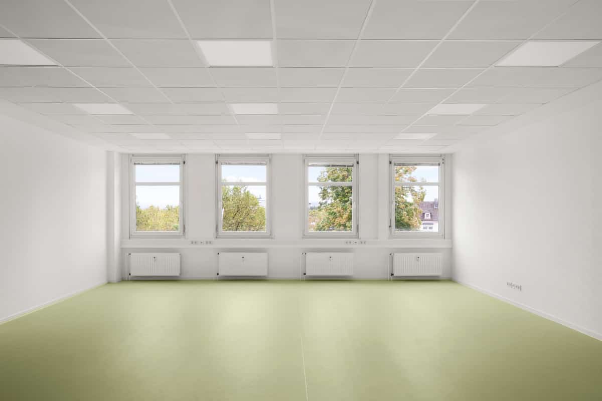 Innenansicht einer leeren Bürofläche mit hellgrünem Bodenbelag, einer Reihe von vier Fenstern, Heizkörpern darunter, weißen Wänden und abgehängter Decke.
