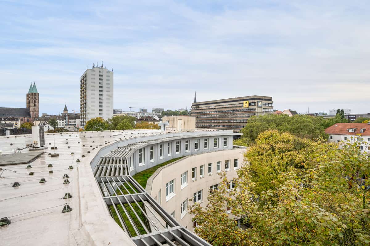 Perspektive vom Dach des Gebäudes auf die geschwungene Gebäudefassade und das begrünte Flachdach, im Hintergrund ein Hochhaus, ein Gebäude mit Baugerüst und gelbem Firmenschild sowie die Kassler Stadtsilhouette mit einem Kirchturm.
