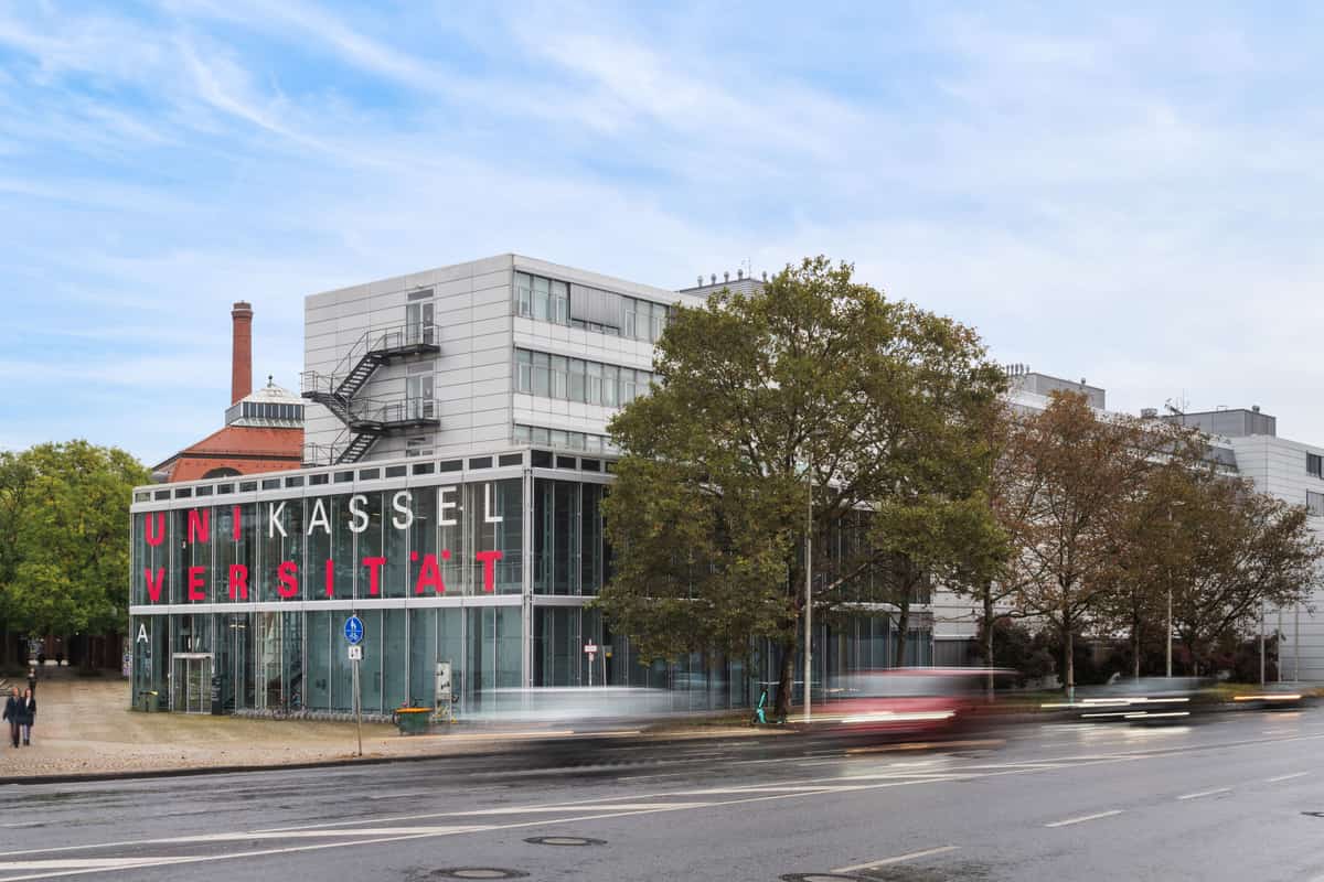 Straßenansicht eines Gebäudes der Universität Kassel in der näheren Umgebung, mit Bewegungsunschärfe durch eine Langzeitbelichtung bei vorbeifahrenden Fahrzeugen. Vereinzelte Fußgänger sind im Hintergrund als unscharfe, nicht erkennbare Silhouetten zu sehen.