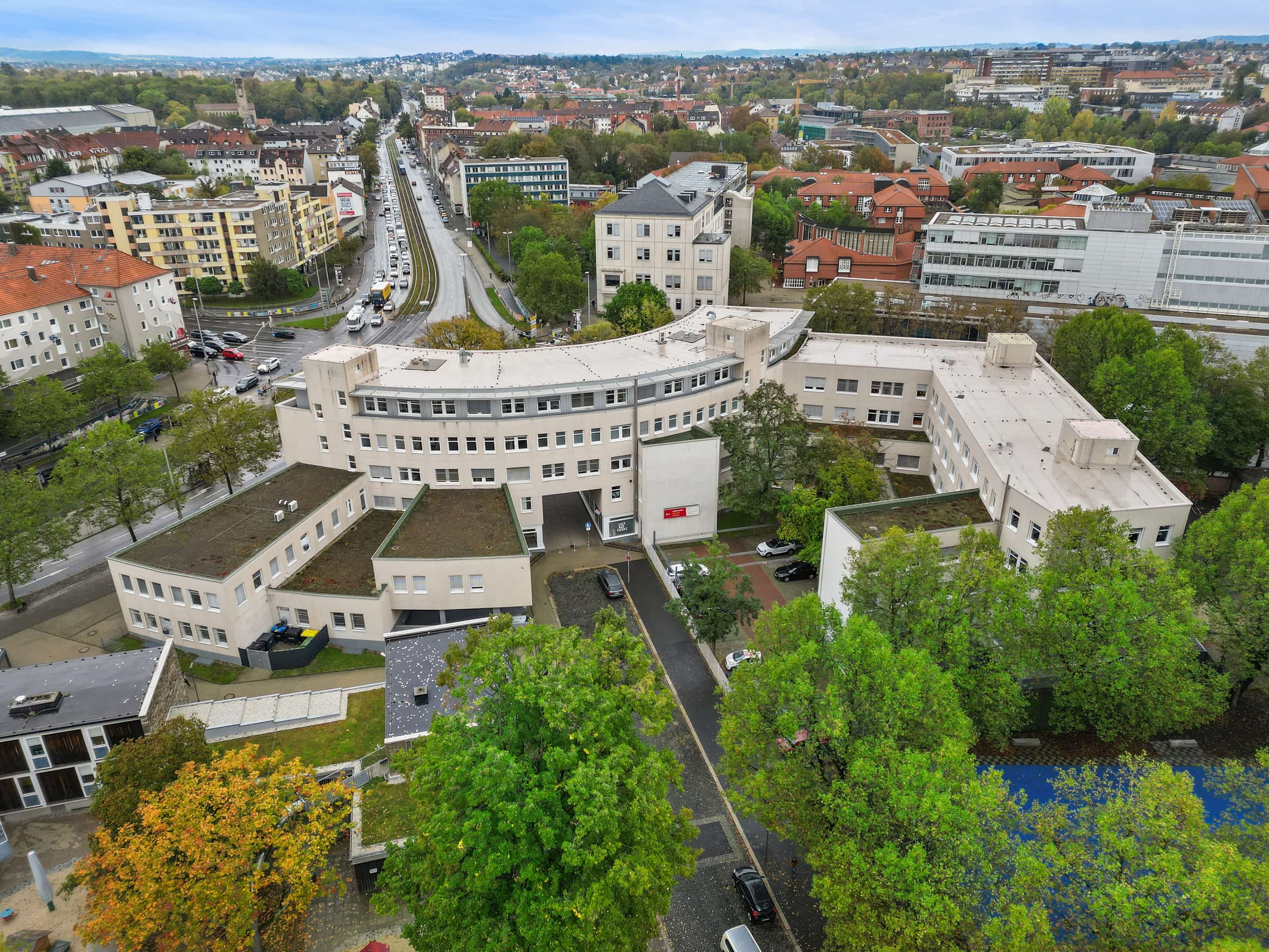 Luftaufnahme aus schräger Perspektive der halbrunden Gewerbeimmobilie am Holländischen Platz in Kassel. Das geschwungene Gebäude mit hellem Fassadenanstrich liegt an einer belebten Straßenkreuzung mit Straßenbahn- und Busverkehr, umgeben von herbstlich gefärbten Bäumen, im Hintergrund die Kassler Stadtsilhouette und bewaldete Hügel.