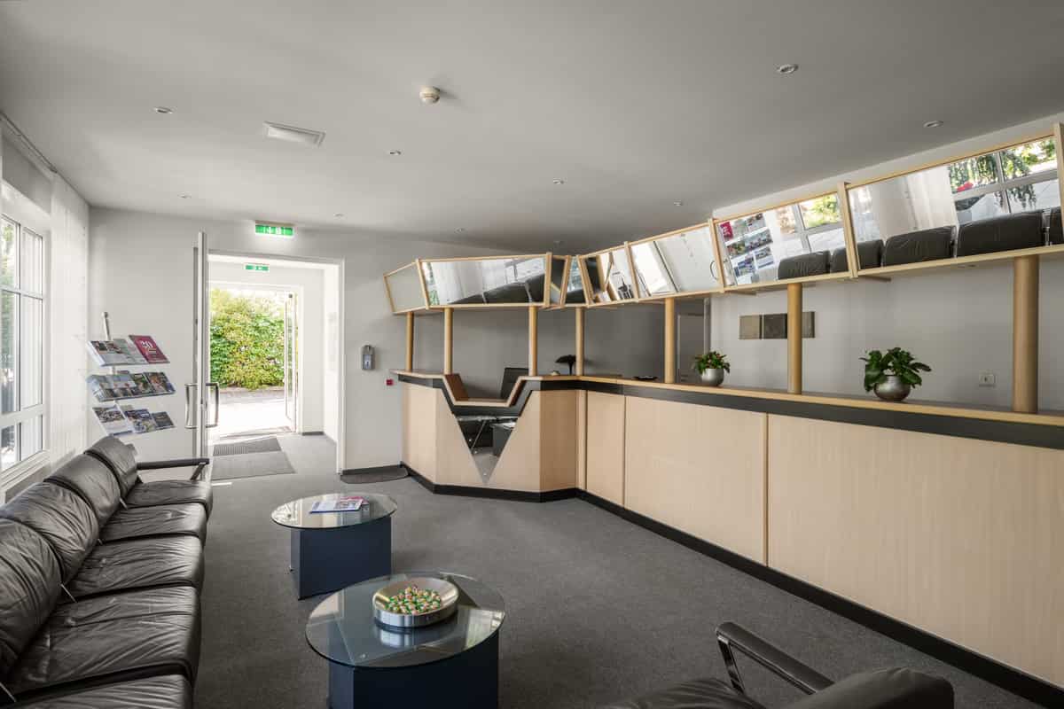 Empfangsbereich mit hellem Holztresen, geometrischer Spiegel-Vordach-Konstruktion, dunklen Ledersofas und Blick durch die geöffnete Eingangstür ins Grüne.