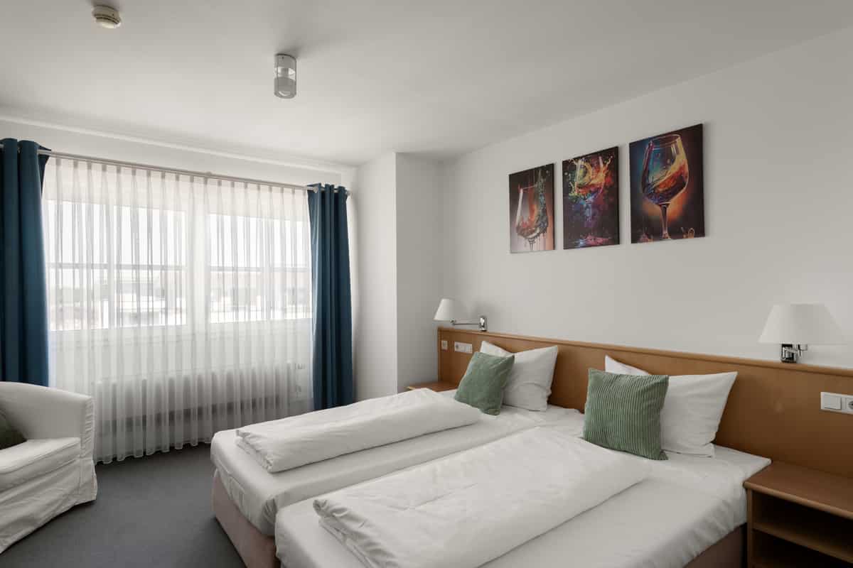 Doppelzimmer mit petrolfarbenen Vorhängen, zwei Betten mit grünen Kissen und einer dreiteiligen Weinglas-Bildserie an der Wand.