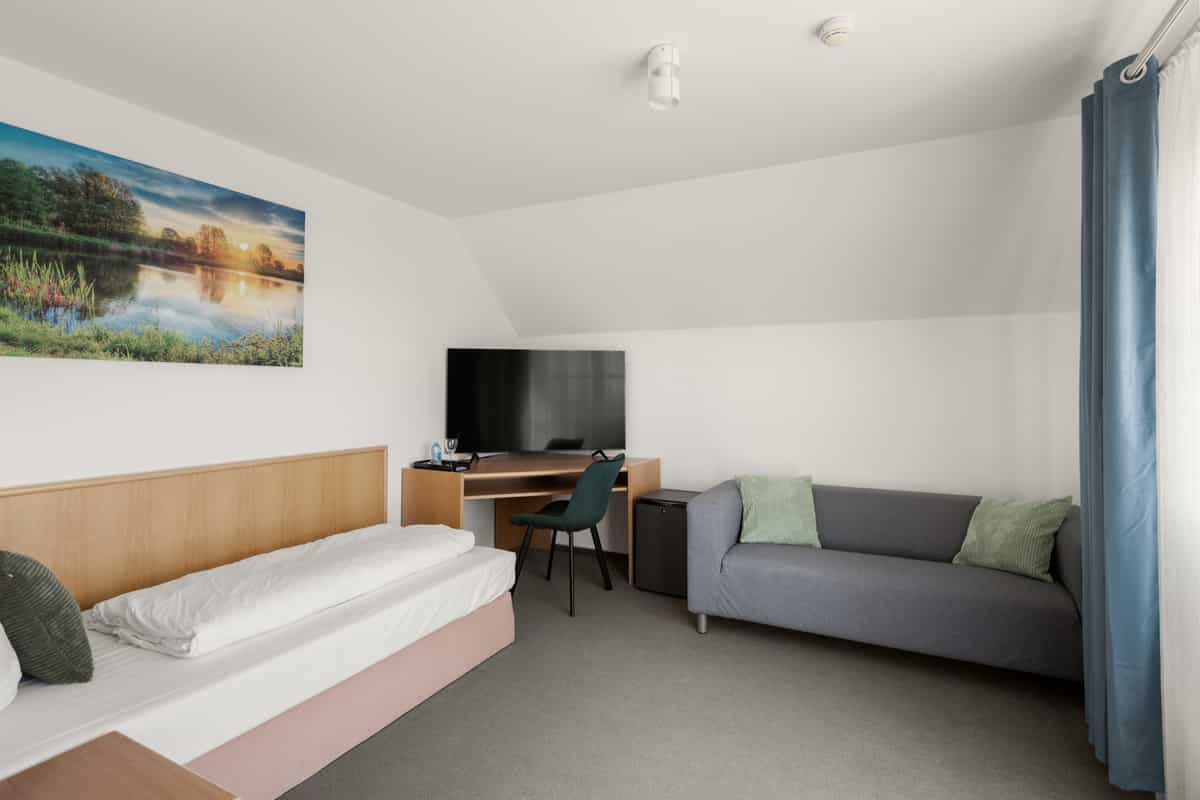 Zimmer mit Einzelbett, grauem Sofa mit grünen Kissen, Schreibtisch mit Fernseher und einem Bild mit Seesonnenuntergang an der Wand.
