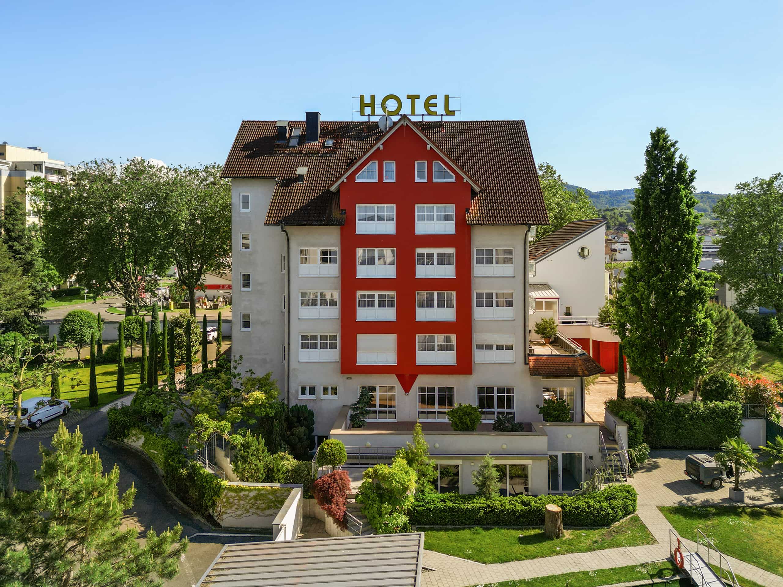 Luftaufnahme des Kirchers Park & Wellnesshotel Kaiserstuhl in Endingen: mehrstöckiges Gebäude mit rotem Giebel, Satteldach und Schriftzug HOTEL, umgeben von Bäumen und Grünflächen.