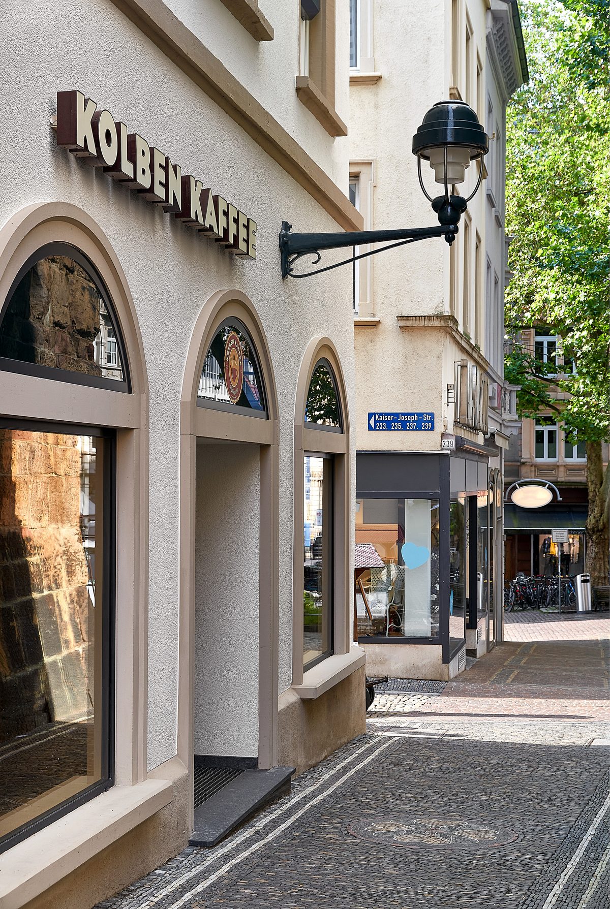 Gebäude mit Kolben Kaffee Schild, gewölbten Fenstern, Laterne und Kopfsteinpflasterweg mit Kanaldeckel.