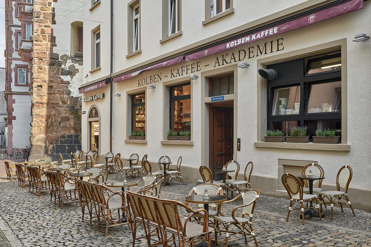 Ein Café mit einer Aufschrift und vielen Tischen und Stühlen auf einem gepflasterten Gehweg.