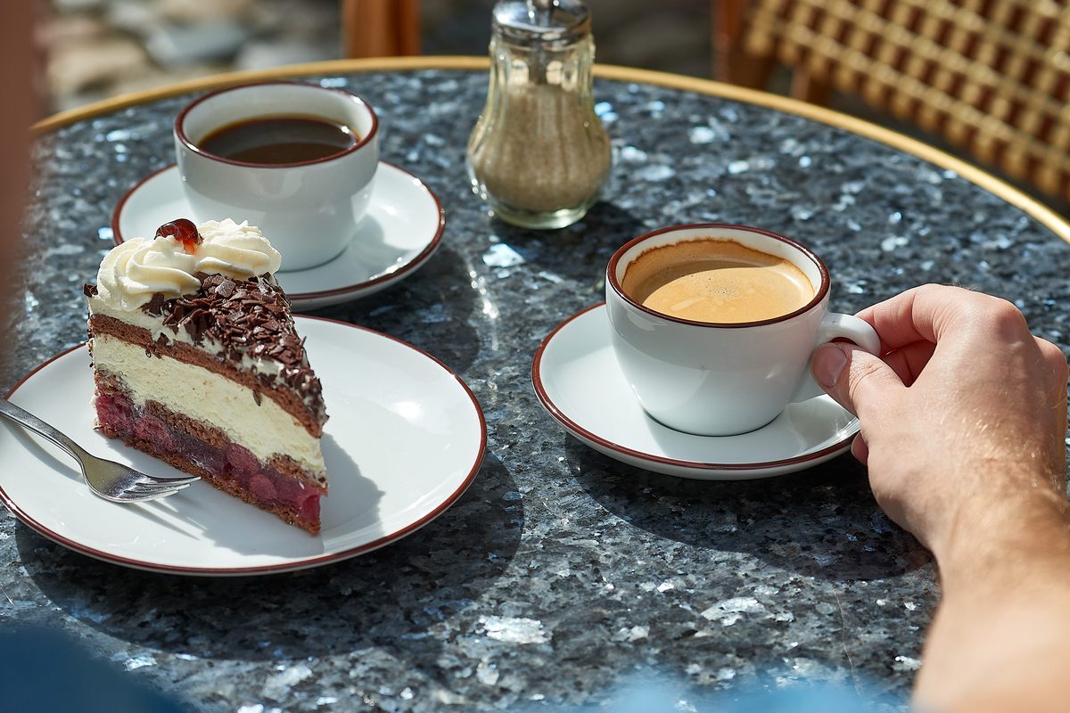 Ein Stück Schwarzwälder Kirschtorte, zwei Tassen Kaffee und ein Zuckerstreuer auf einem dunklen Steintisch.