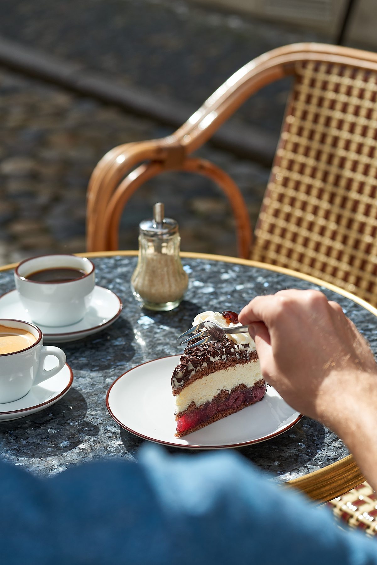Eine Hand isst ein Stück Schwarzwälder Kirschtorte mit einer Gabel an einem Tisch mit zwei Kaffeetassen und Zuckerstreuer.
