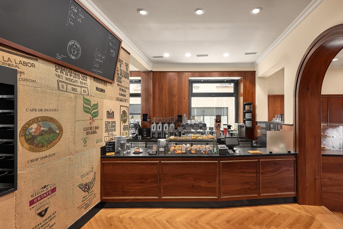 Eine Theke mit Gebäckauslage und Kaffeemaschinen in einem Café mit Holzboden und Wänden aus Kaffeesäcken.