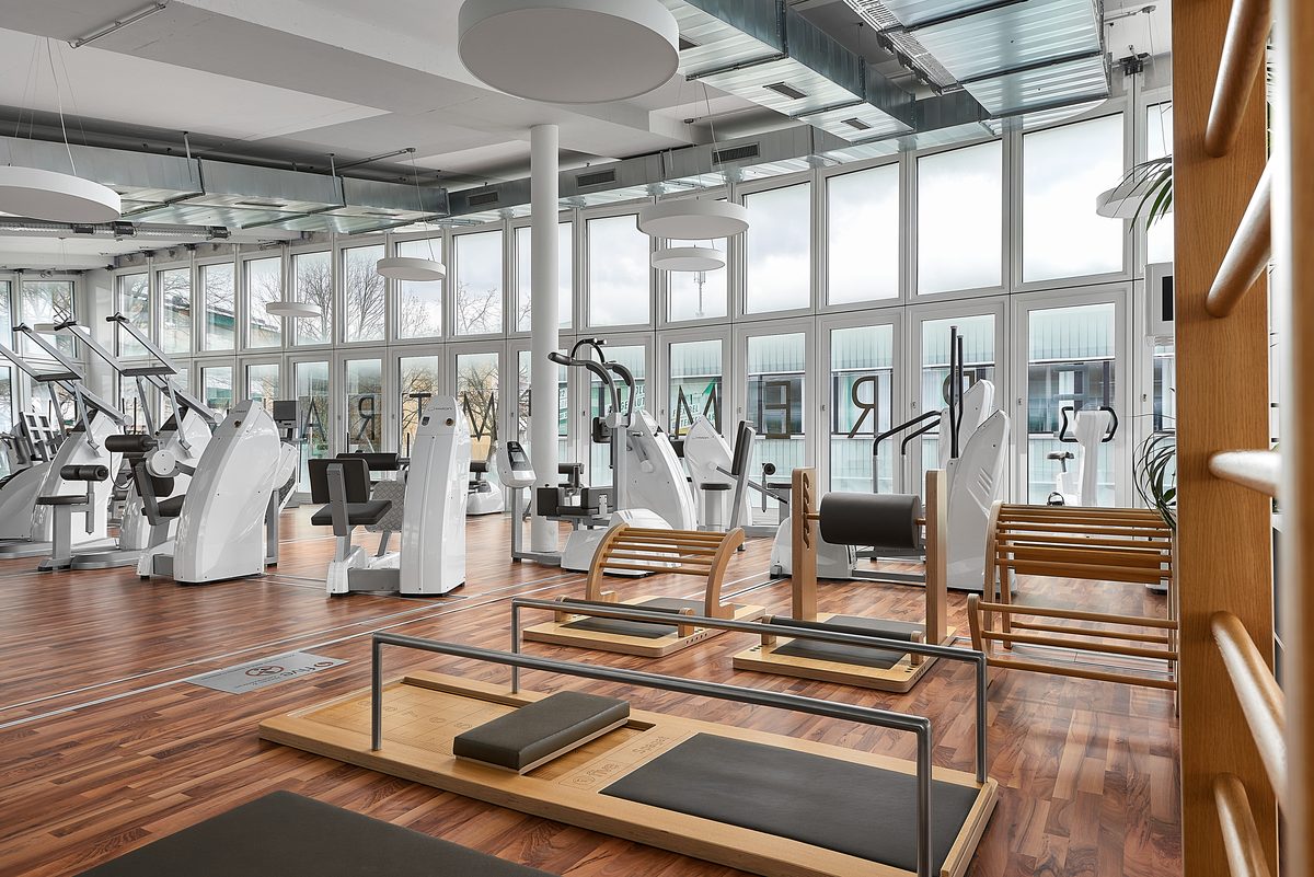 Ein Fitnessstudio mit Holzboden, grossen Fenstern, weissen Trainingsgeraeten und Holz-Trainingsbänken.