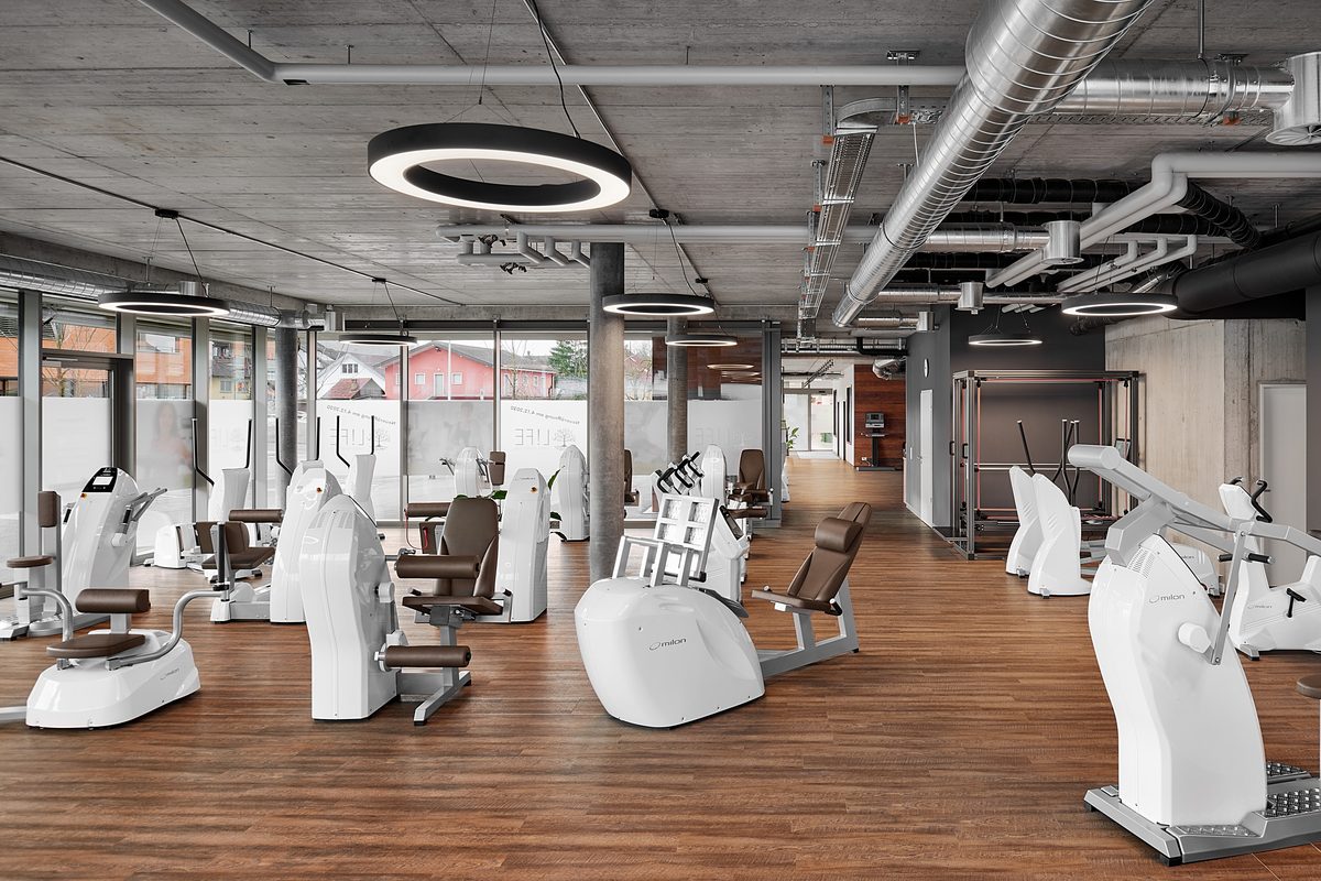 Ein Fitnessstudio mit Holzboden, grauer Decke, runden Lampen und mehreren weißen Trainingsgeräten.