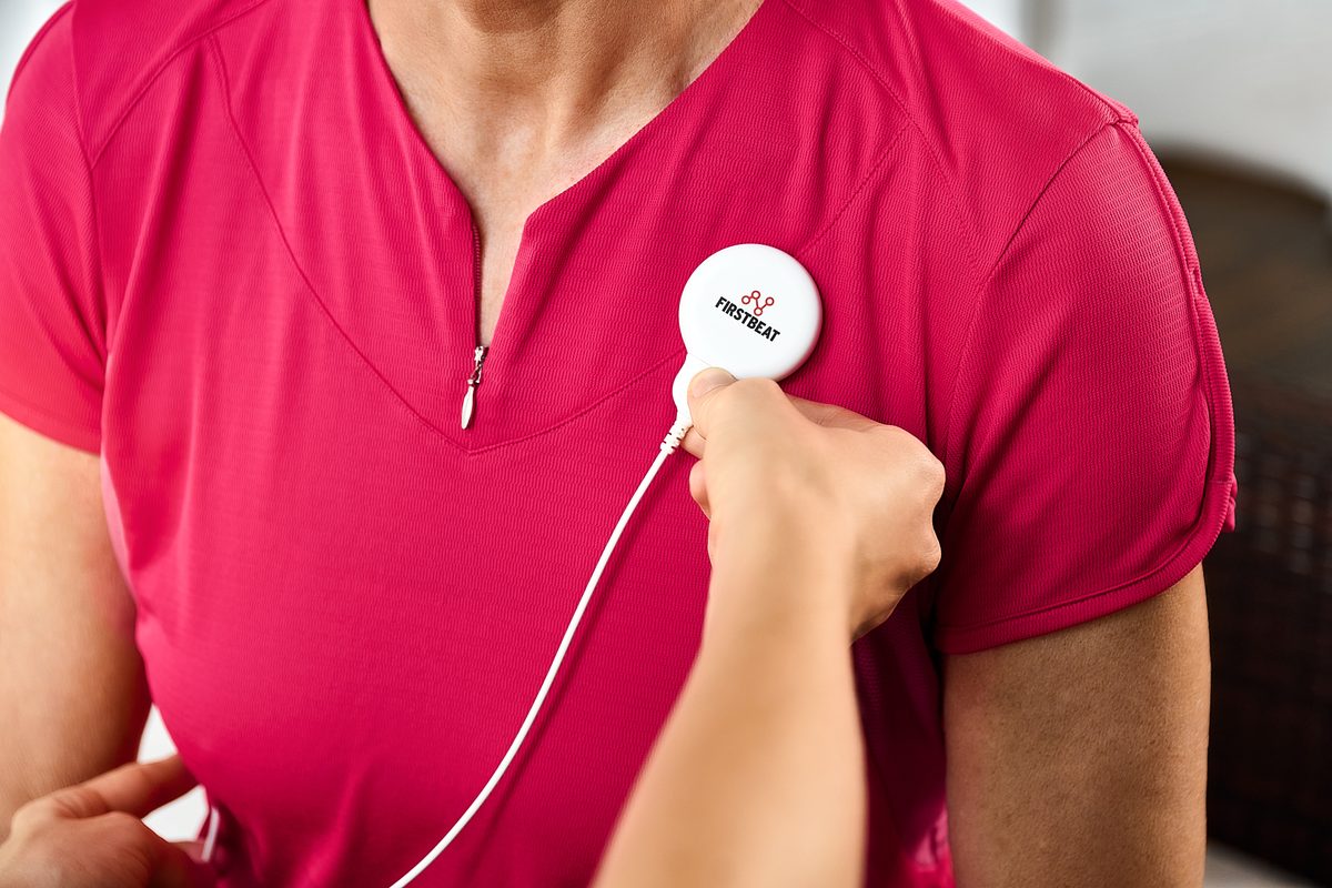 Eine Person in einem pinken Sportshirt befestigt ein weißes Firstbeat-Gerät mit Kabel an ihrer Brust.