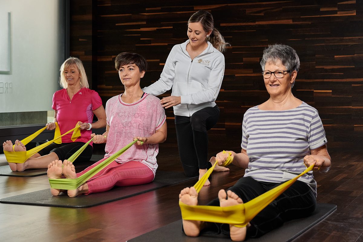 Drei Frauen trainieren mit Fitnessbändern auf Matten, eine Trainerin korrigiert die Haltung einer Teilnehmerin.