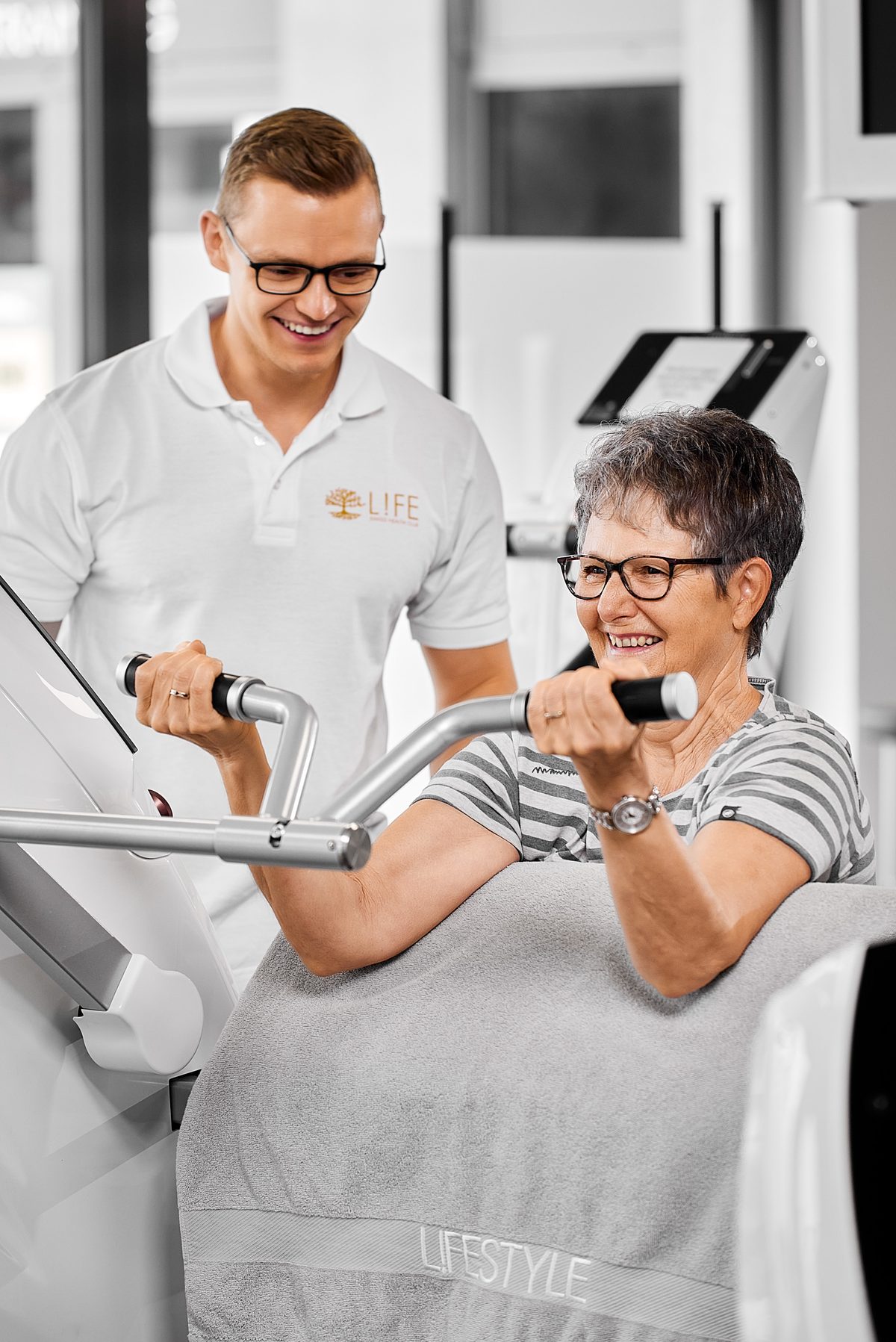 Ein Trainer in einem weißen Poloshirt lächelt einer lächelnden Frau mit Brille zu, die an einem Fitnessgerät trainiert.