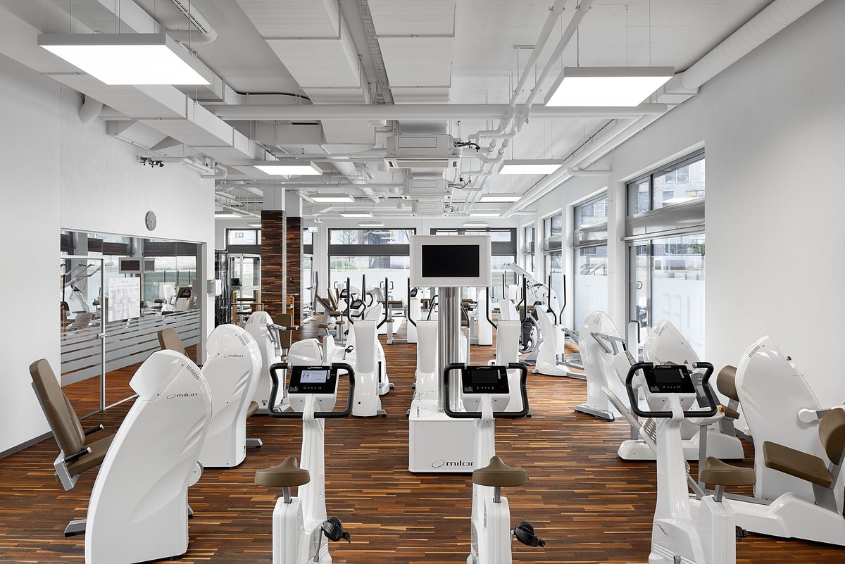 Ein Fitnessstudio mit Holzboden, weißen Wänden und Decken, sowie zahlreichen weißen Trainingsgeräten und großen Fenstern.