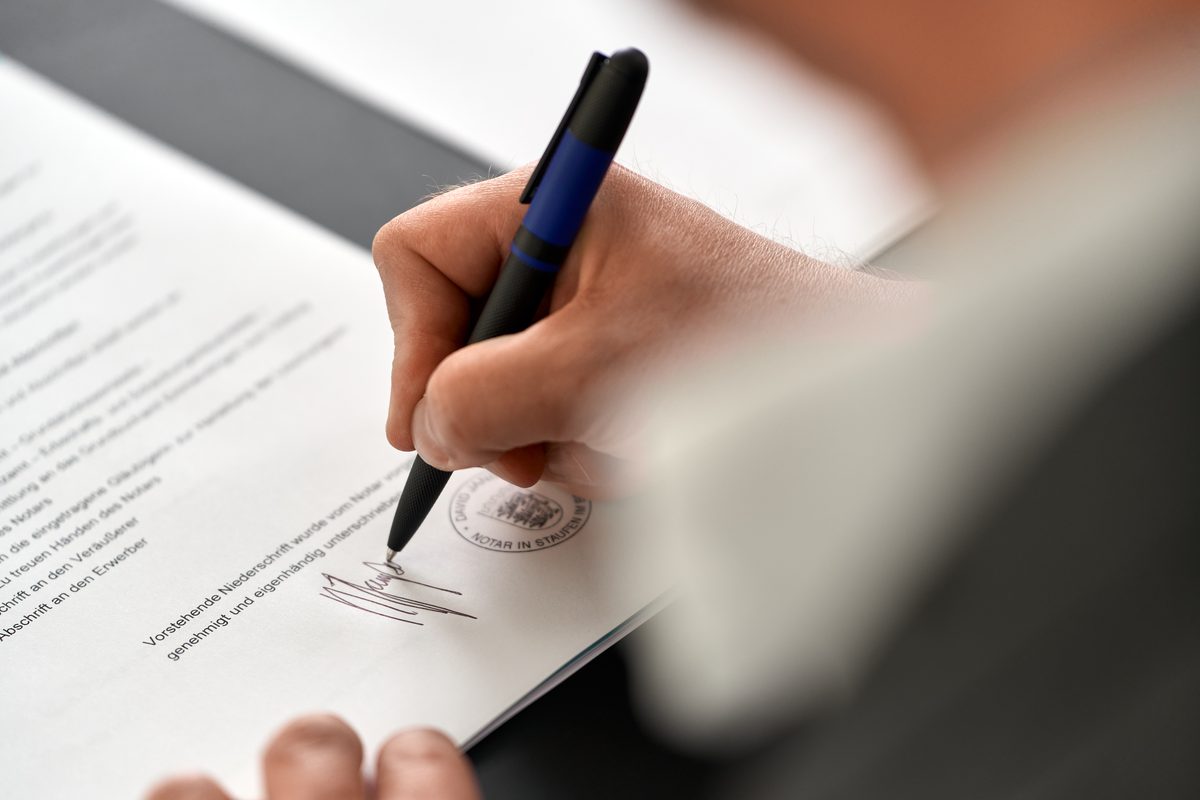 Eine Hand unterschreibt mit einem schwarzen Kugelschreiber ein Dokument mit einem Stempel.
