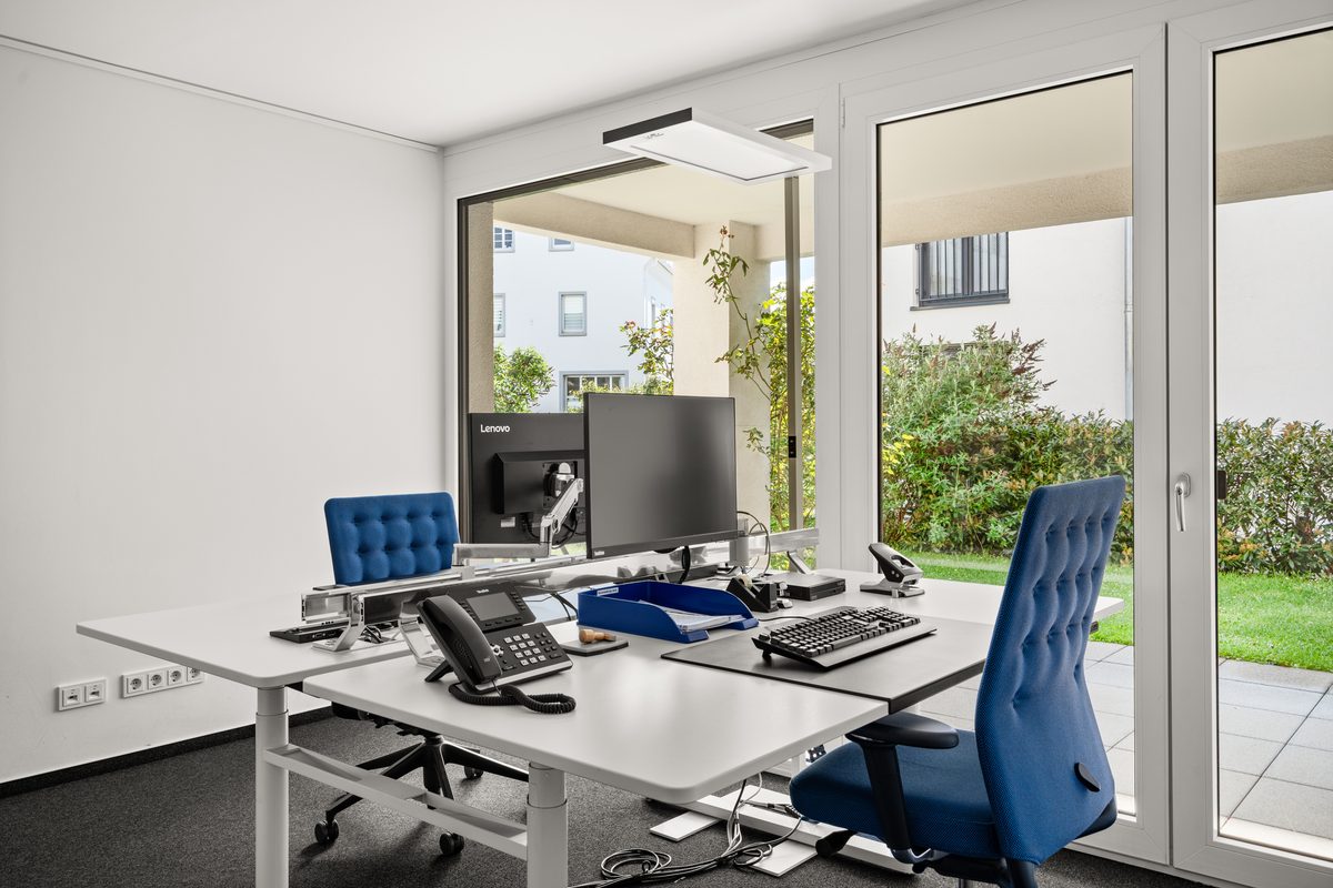 Ein moderner Büroraum mit zwei Schreibtischen, blauen Bürostühlen und Computern vor einer großen Fensterfront mit Blick ins Grüne.