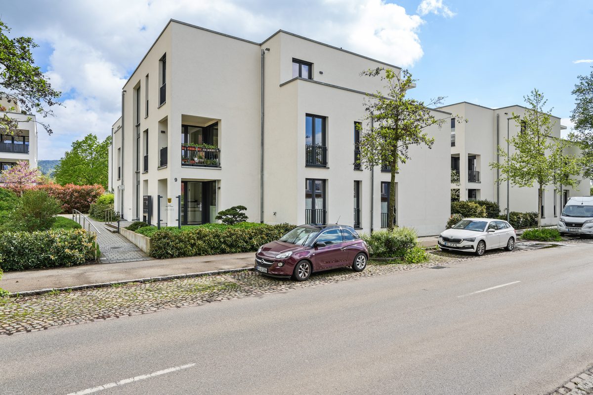 Mehrere weiße Gebäude mit Balkonen und Fenstern, davor parken Autos am Straßenrand, umgeben von Bäumen und Büschen.