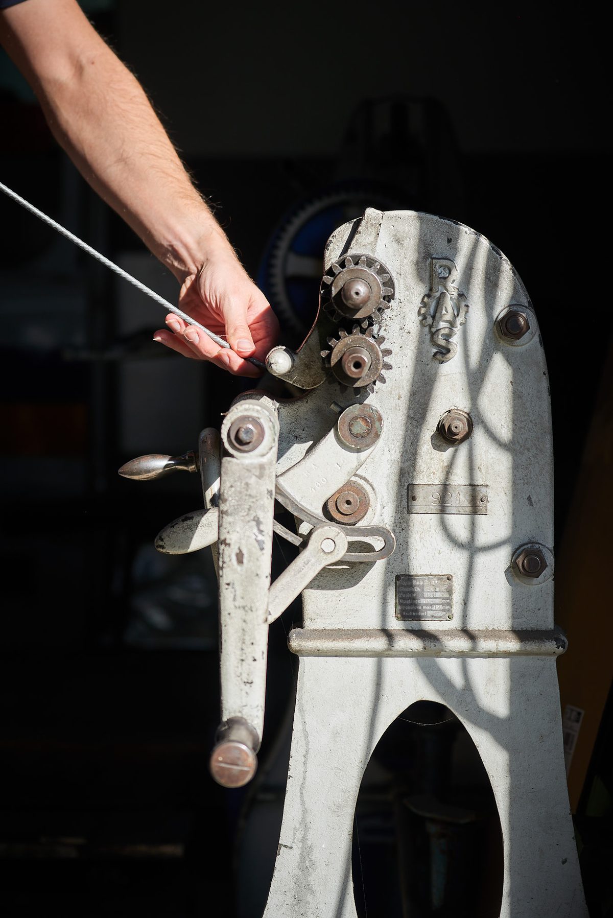 Ein Arm führt ein Metallkabel in eine alte, graue Maschine mit Zahnrädern und Hebeln, die im Sonnenlicht steht.