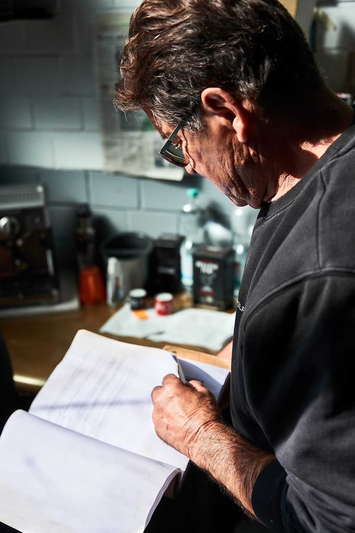 Ein Mann mit Brille und dunklem Pullover blättert in einem großen Buch, im Hintergrund eine Theke mit Geräten.