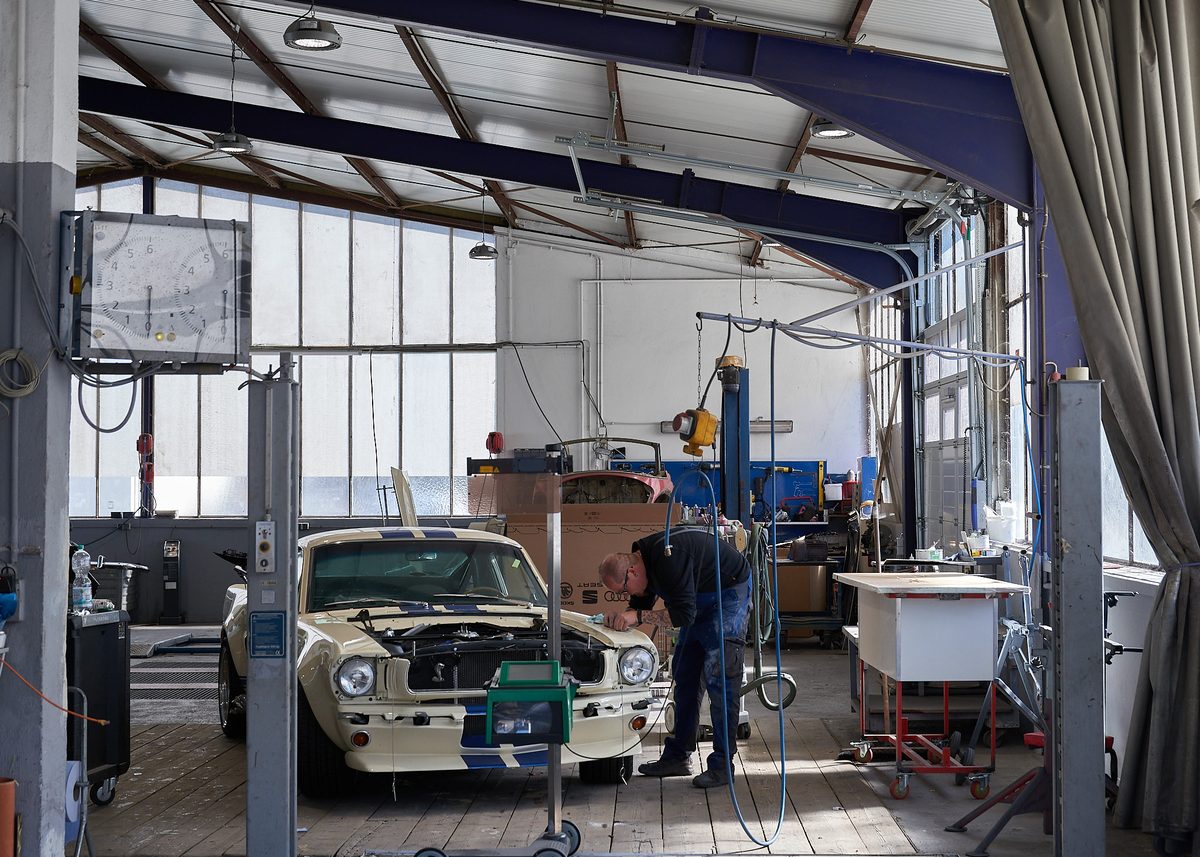Ein Mann arbeitet an einem beigen Oldtimer mit blauen Streifen in einer Werkstatt mit großen Fenstern.