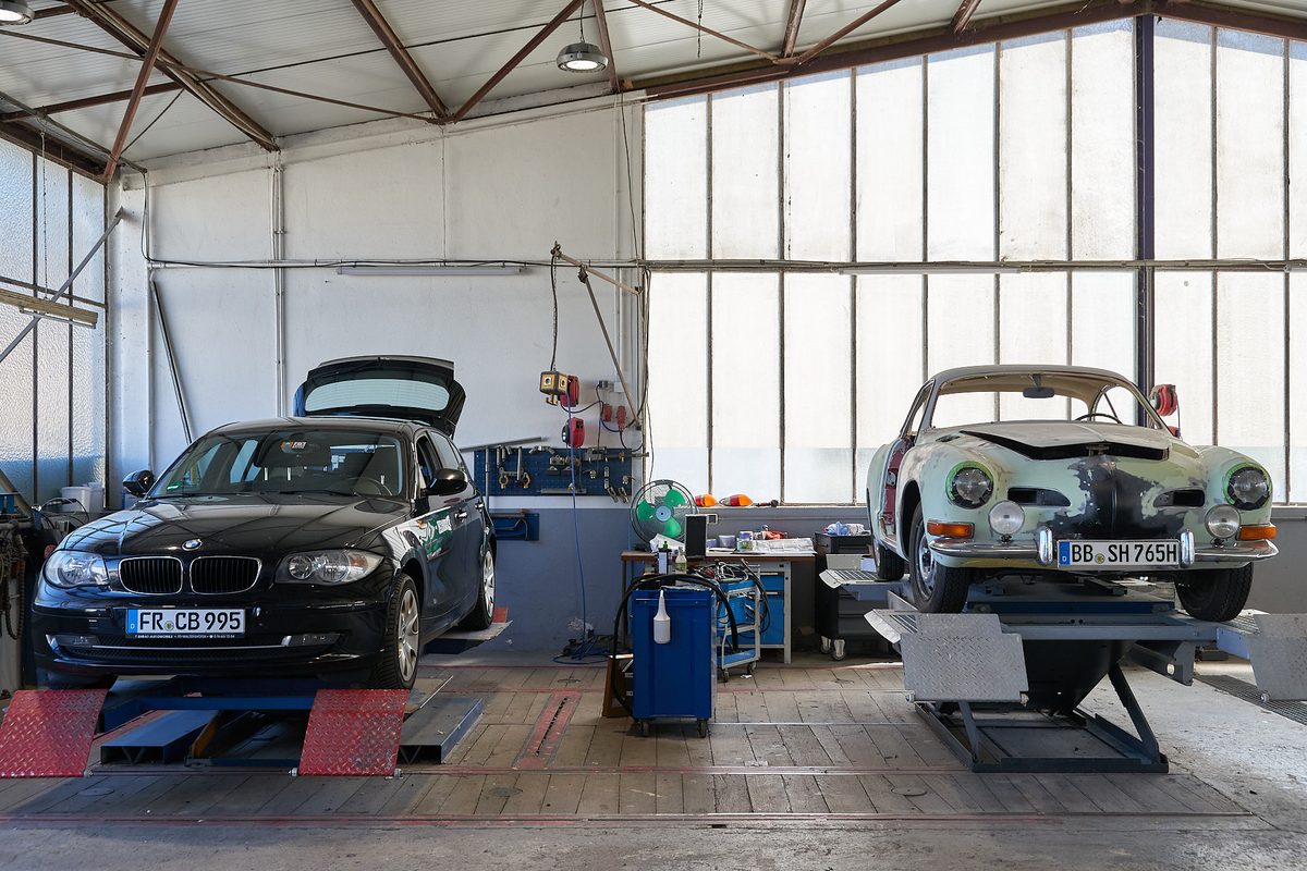 Zwei Autos stehen in einer Werkstatt auf Hebebühnen, ein schwarzer BMW und ein hellgrüner Oldtimer.