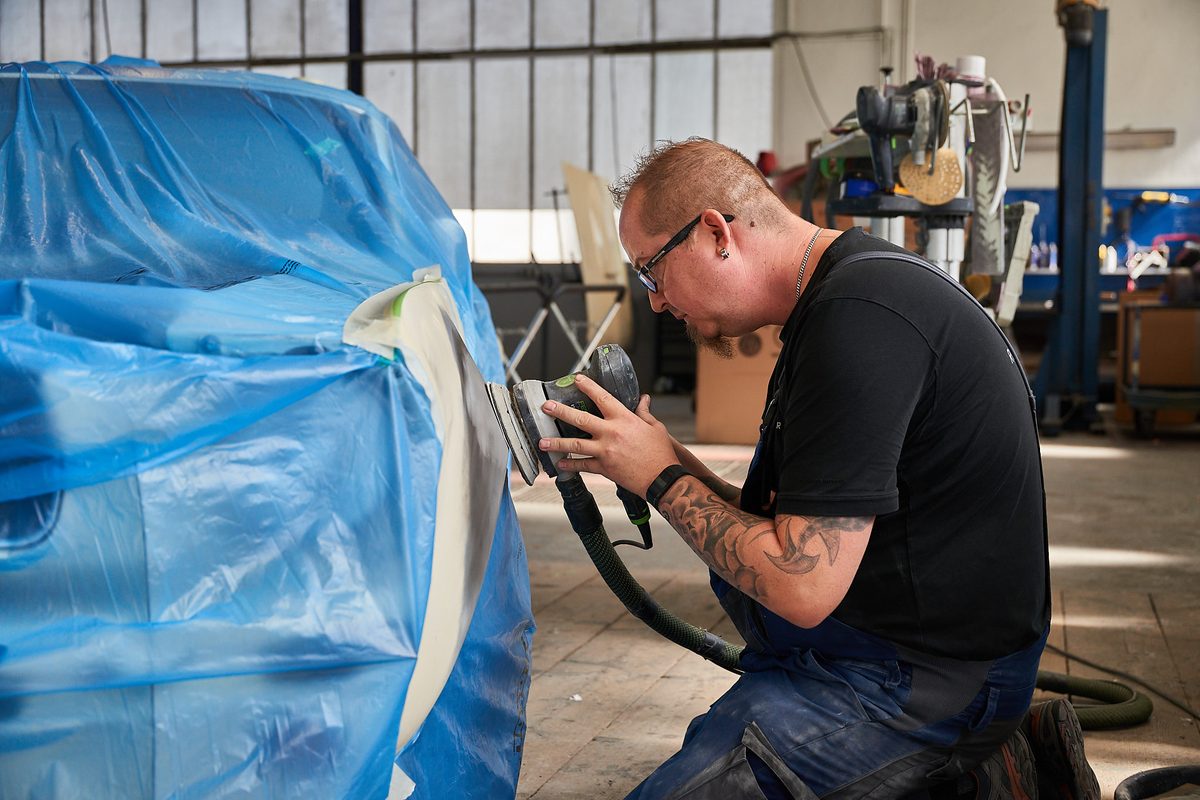 Ein Mann kniet und schleift mit einer Maschine die Karosserie eines blauen, abgedeckten Autos in einer Werkstatt.