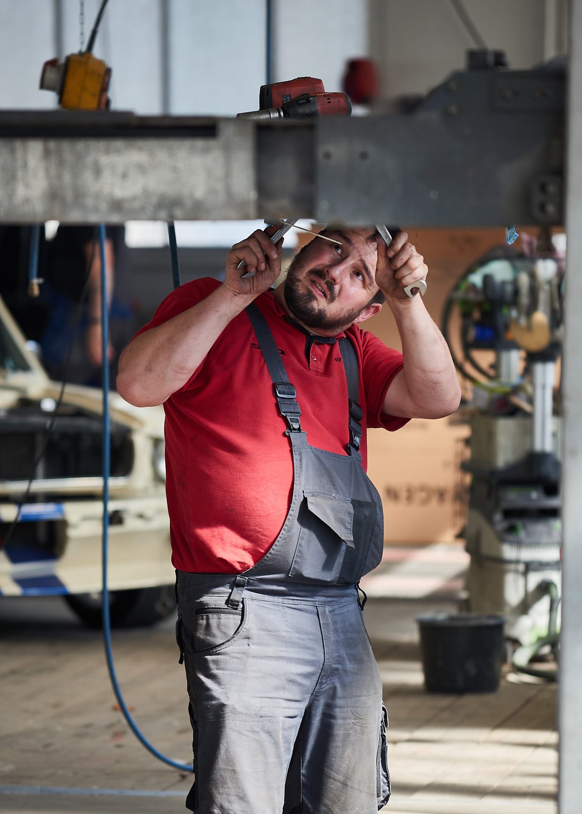 Ein Mann in rotem T-Shirt und grauer Latzhose repariert mit zwei Schraubenschlüsseln ein Bauteil über seinem Kopf.