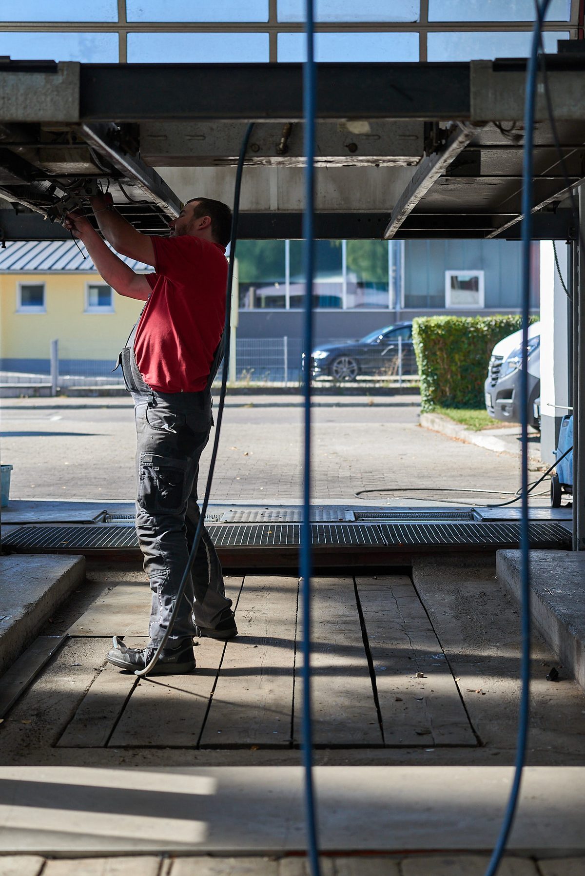 Ein Mann in rotem T-Shirt und Arbeitshose arbeitet unter einem Fahrzeug auf einer Hebebühne in einer Werkstatt.