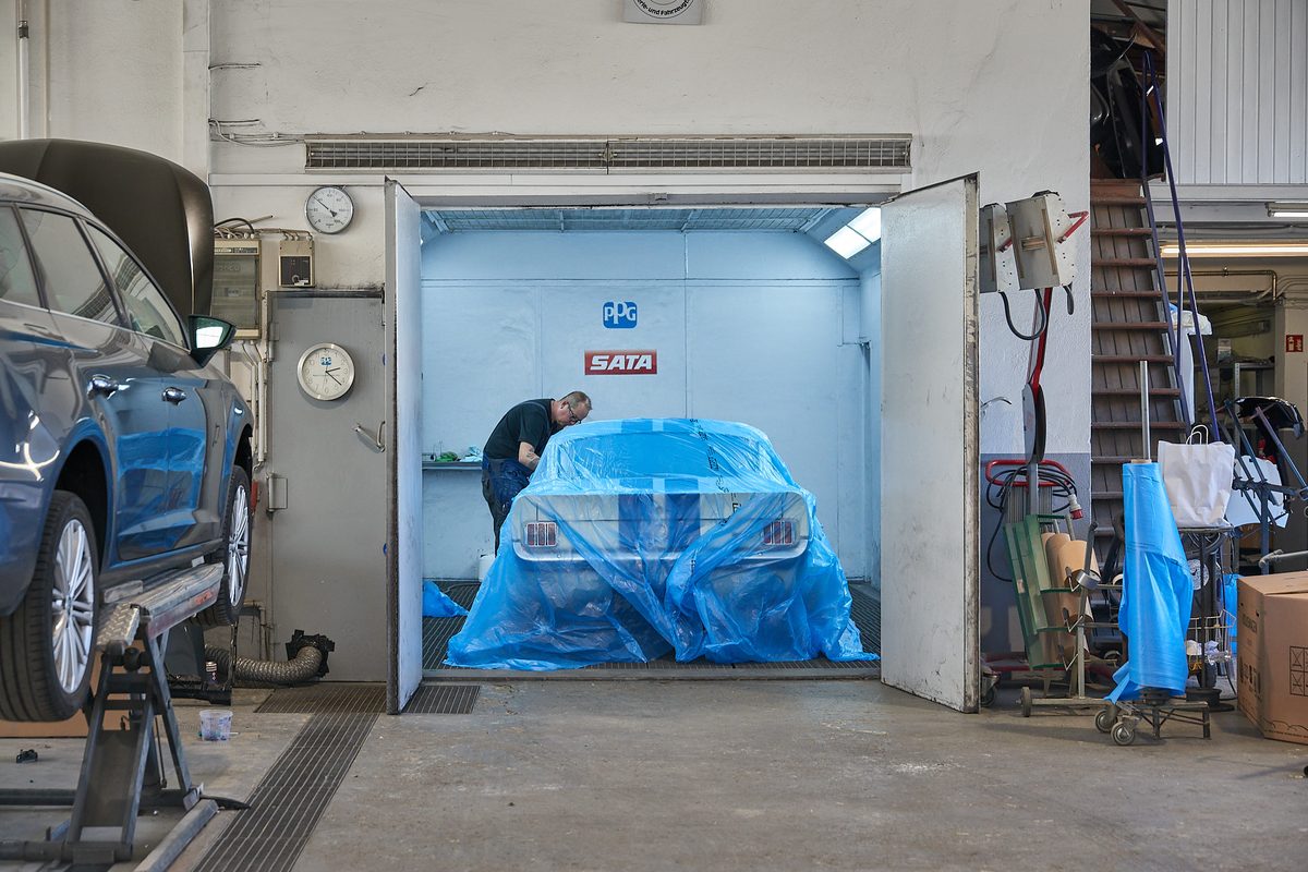 Ein Mann arbeitet in einer Lackierkabine an einem teilweise abgedeckten Auto, während ein anderes Auto auf einer Hebebühne steht.
