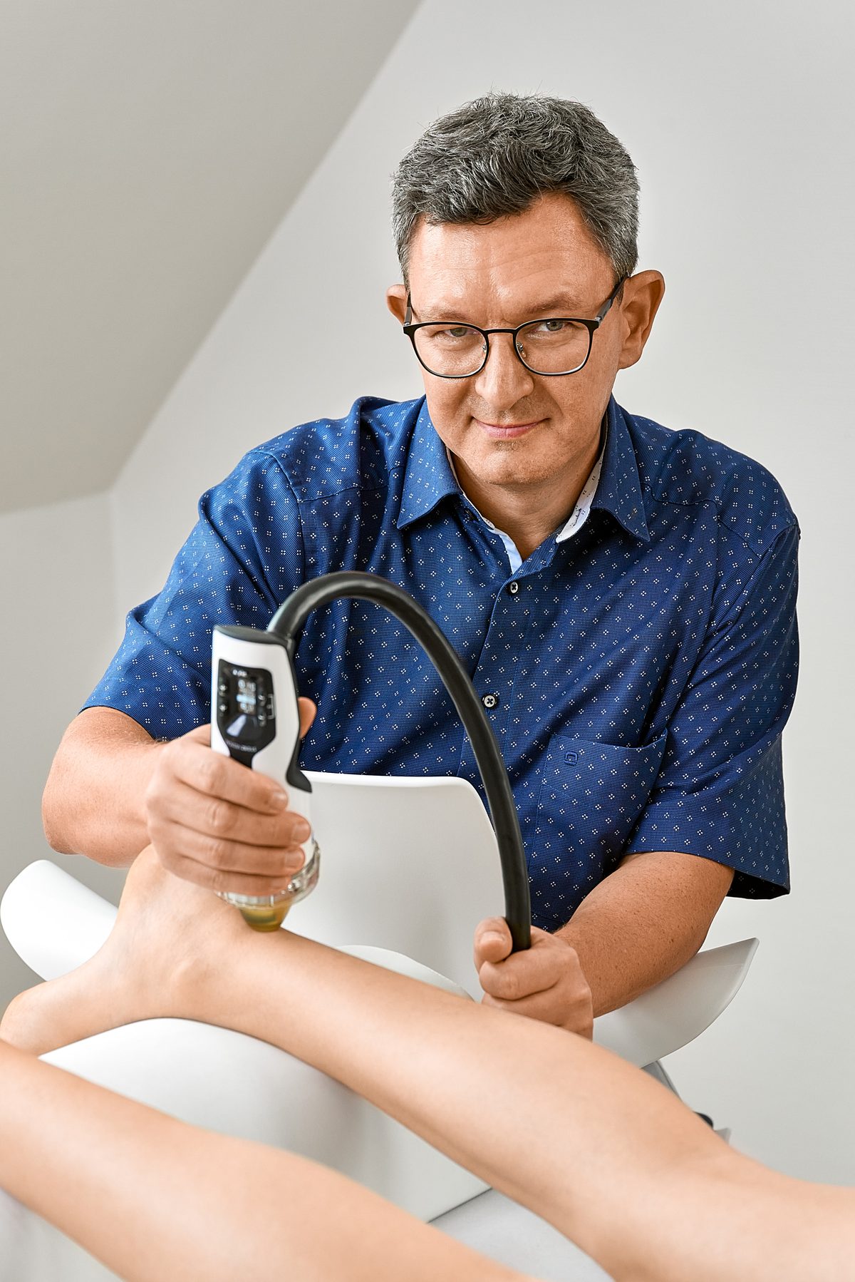 Ein Mann mit Brille und blauem Hemd behandelt den Fuß einer Person mit einem medizinischen Gerät.