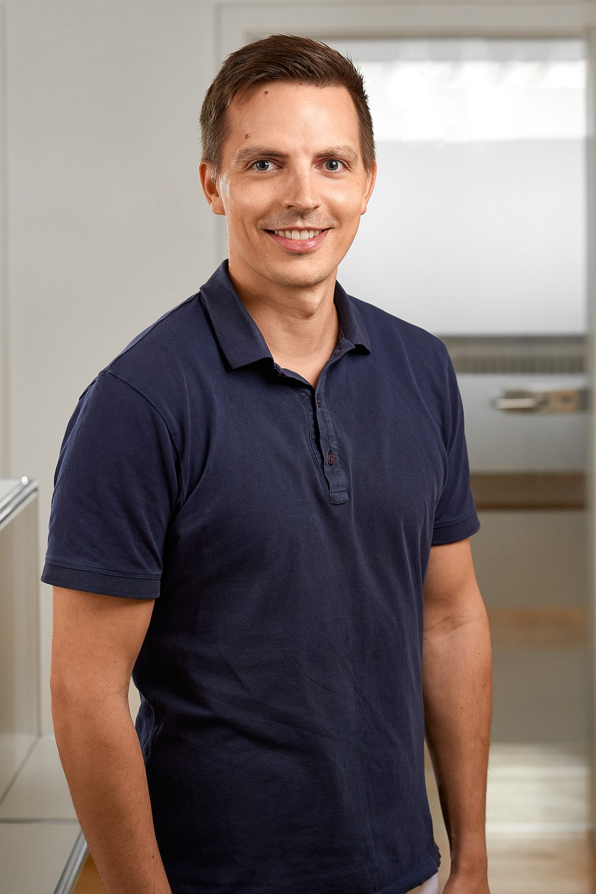 Ein lächelnder Mann in einem dunkelblauen Poloshirt steht vor einem hellen Hintergrund.