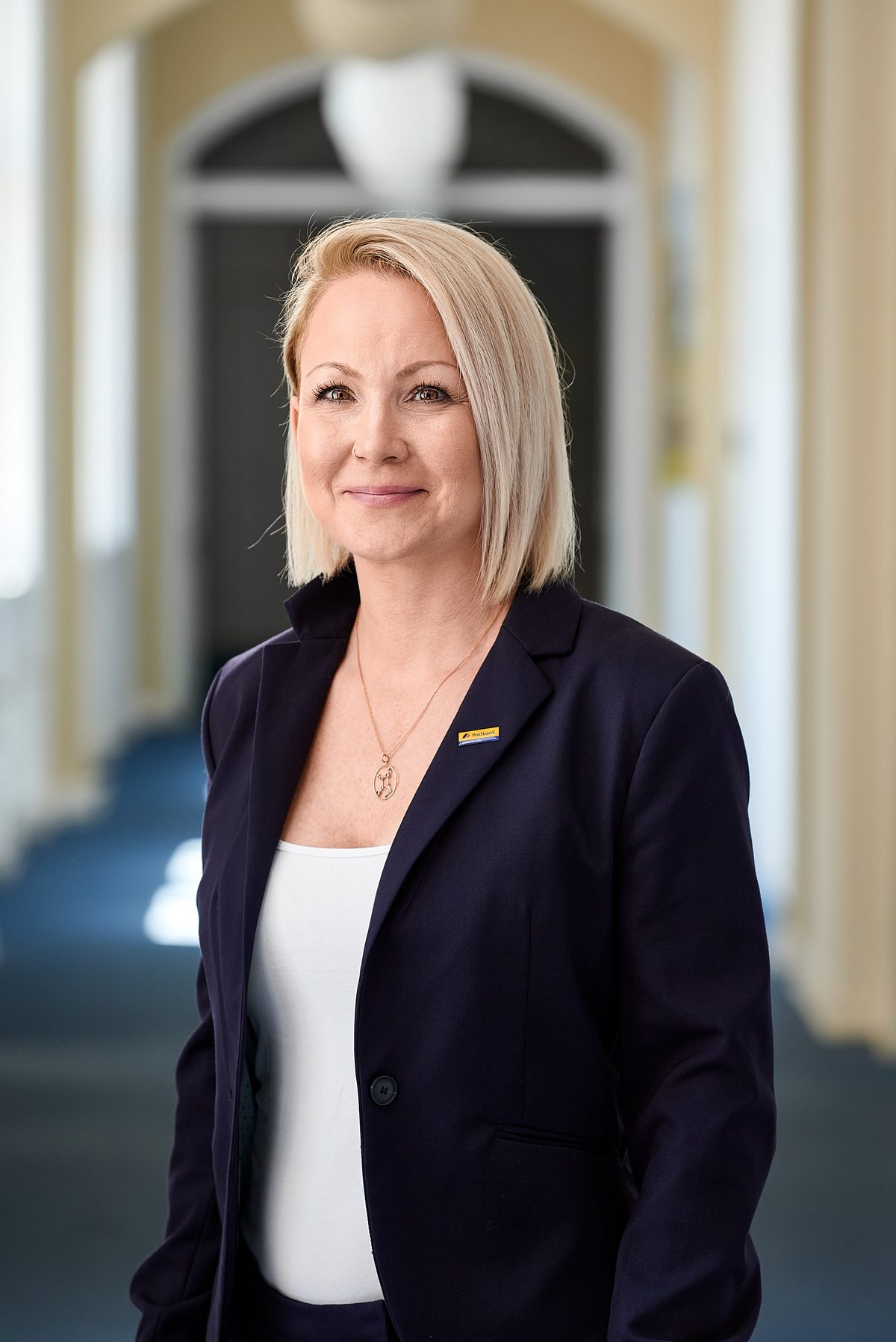 Eine blonde Frau mit dunklem Blazer und weißem Oberteil steht lächelnd in einem hellen Flur.