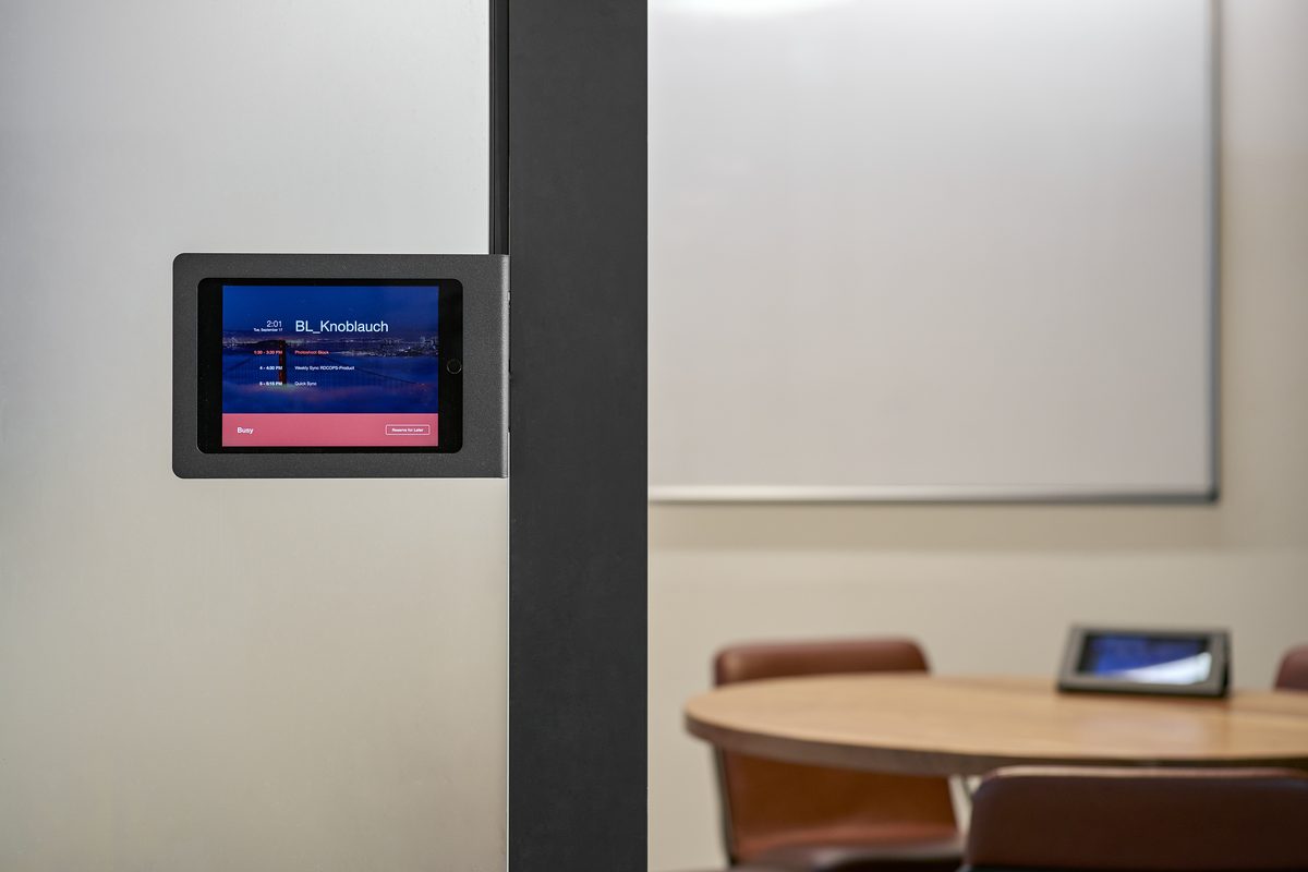 Ein Tablet mit Terminkalender ist an einer Wand befestigt, im Hintergrund ein Konferenztisch mit Stühlen und eine weiße Tafel.