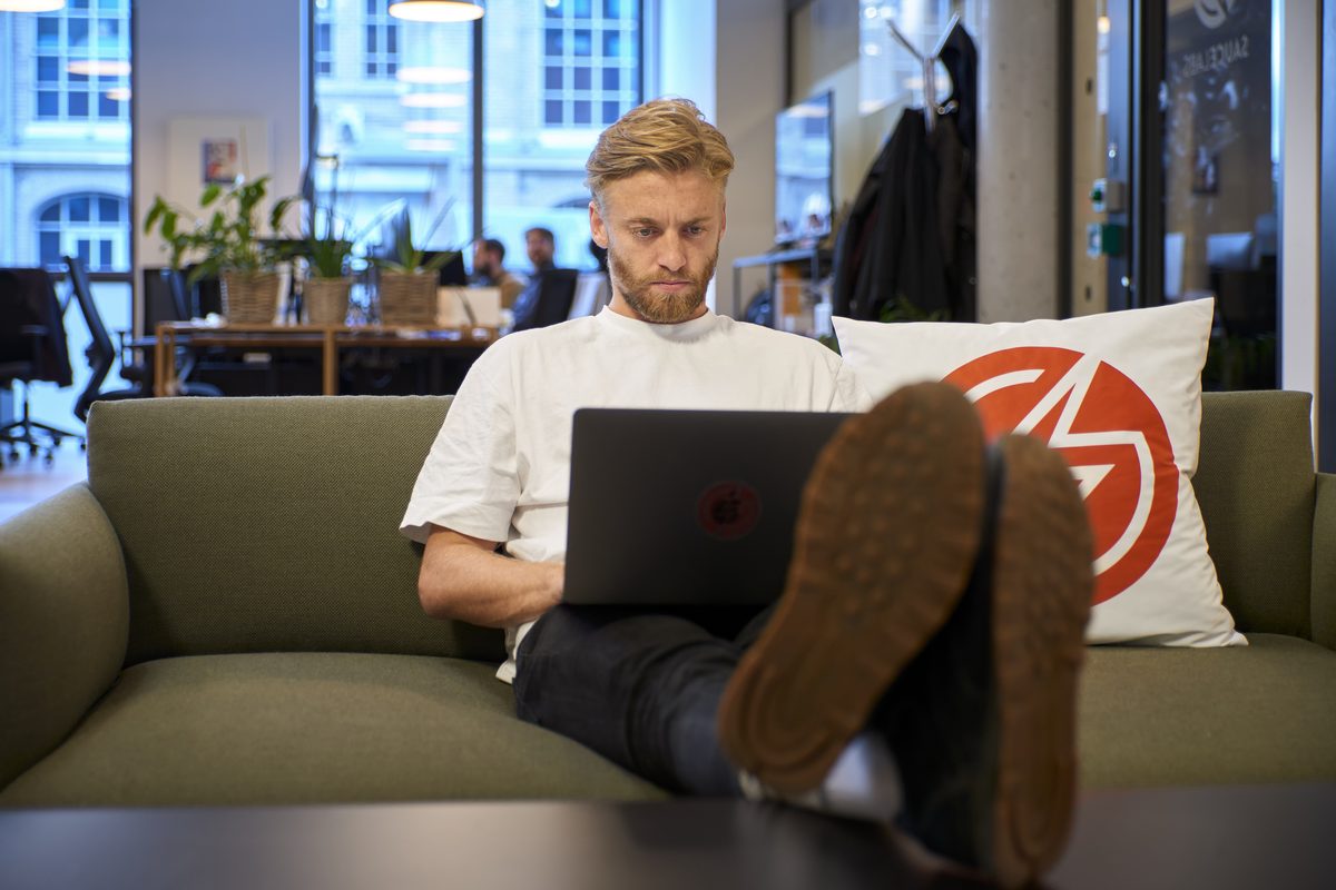 Ein Mann mit Bart sitzt auf einem Sofa, die Füße auf einem Tisch, und arbeitet an einem Laptop.