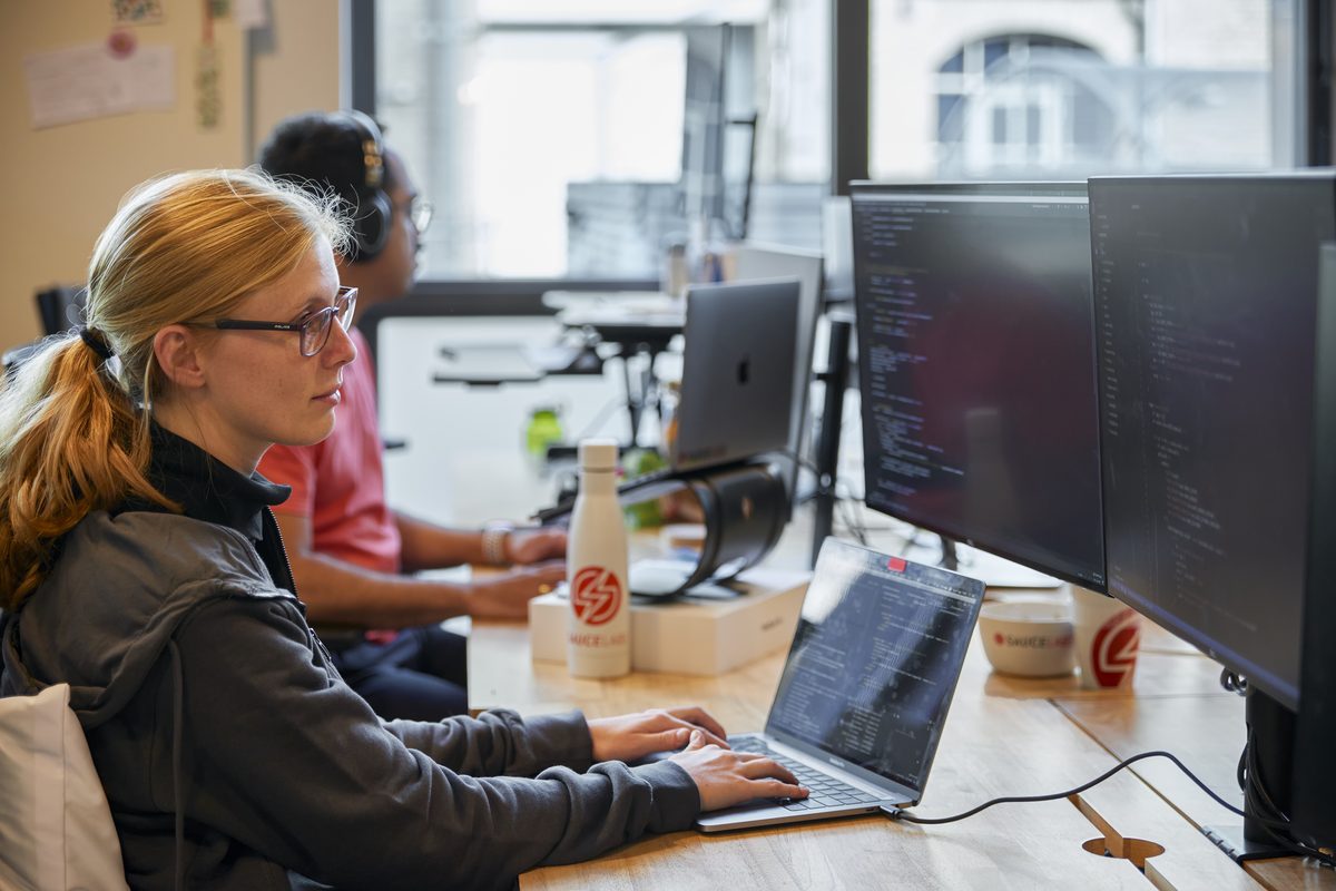 Eine Frau mit Brille und Zopf tippt an einem Schreibtisch auf einem Laptop, daneben zwei Monitore mit Code.