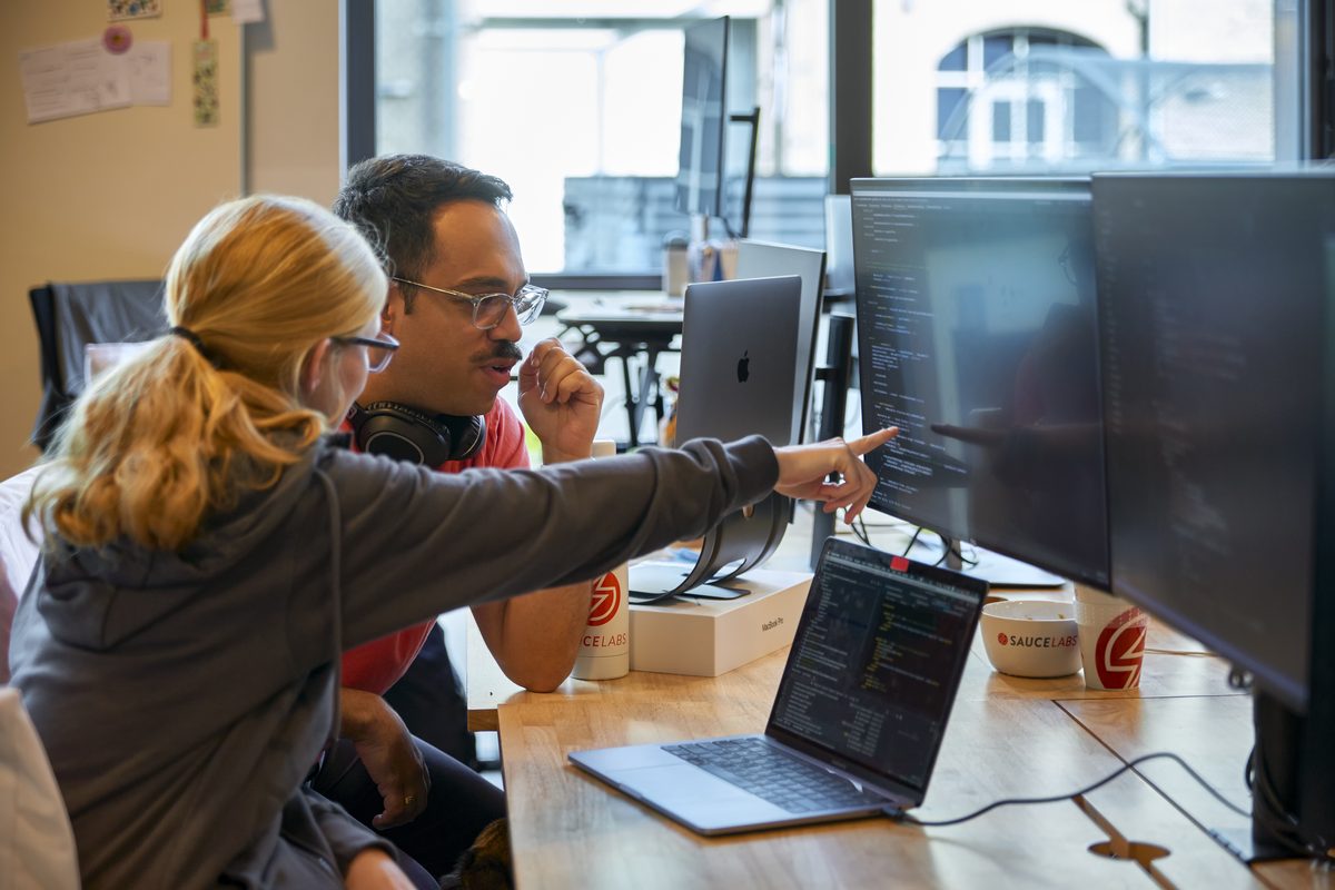 Zwei Personen sitzen an einem Holztisch und schauen auf einen Computermonitor mit Code, eine Person zeigt auf den Bildschirm.